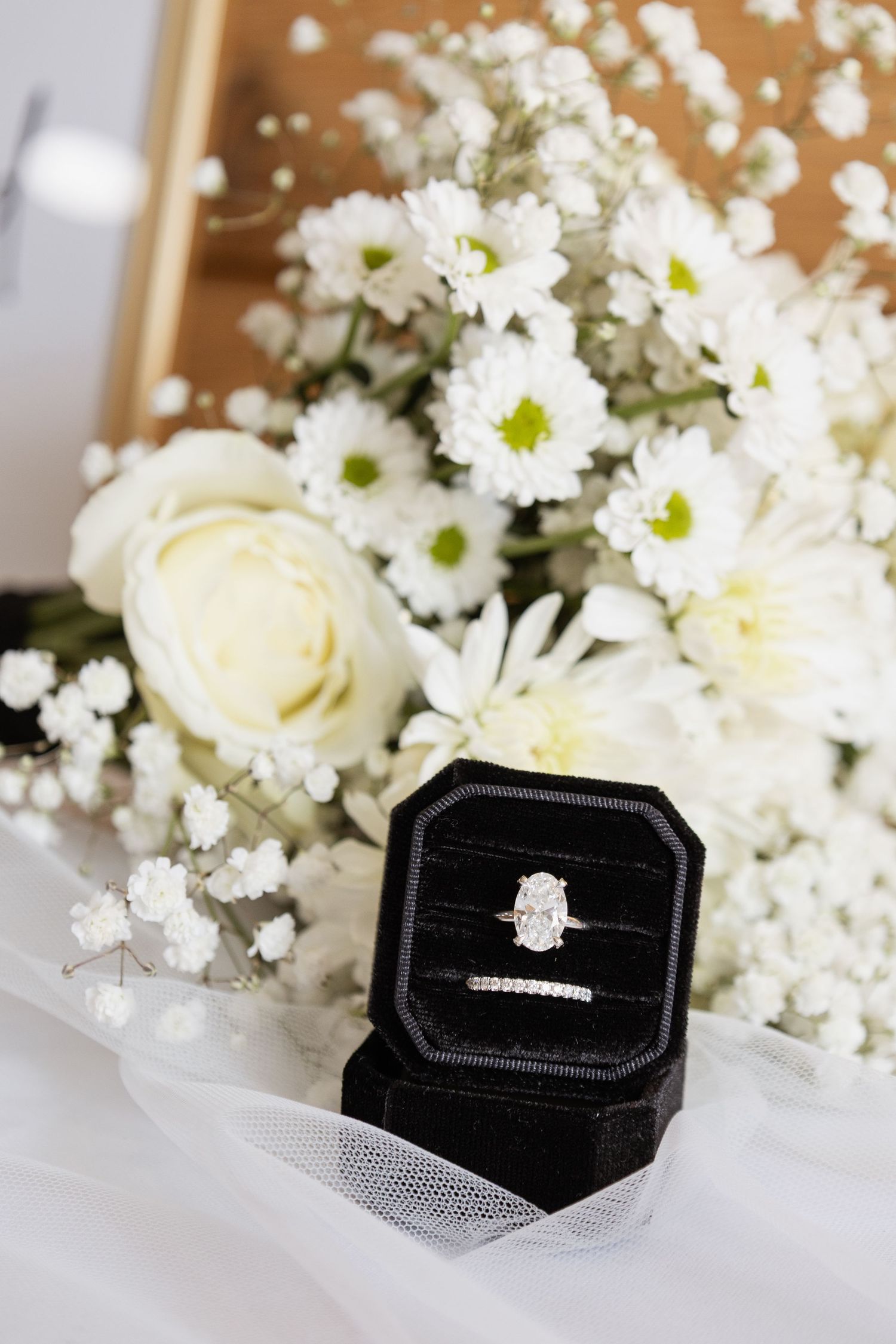 Diamond engagement ring in black velvet box with white roses and daisies bouquet wedding detail photo.