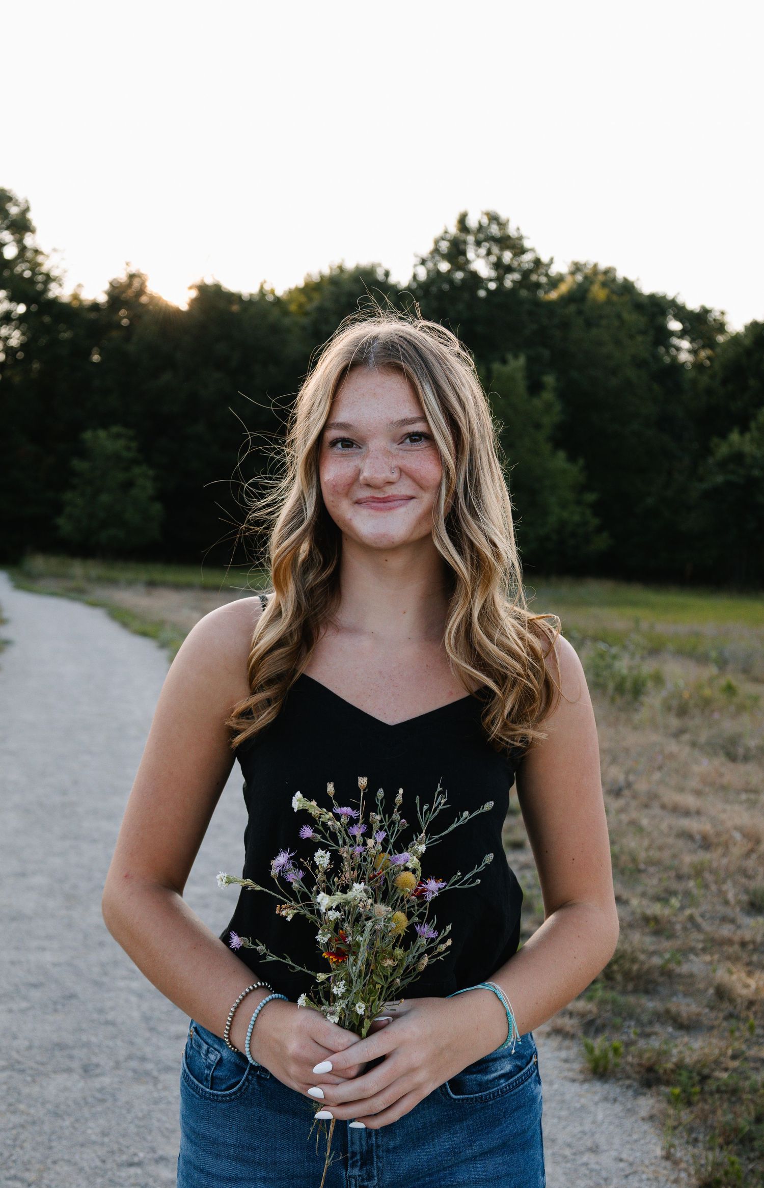 Young woman with wavy blonde hair smiles while holding wildflowers on a scenic path at golden hour.