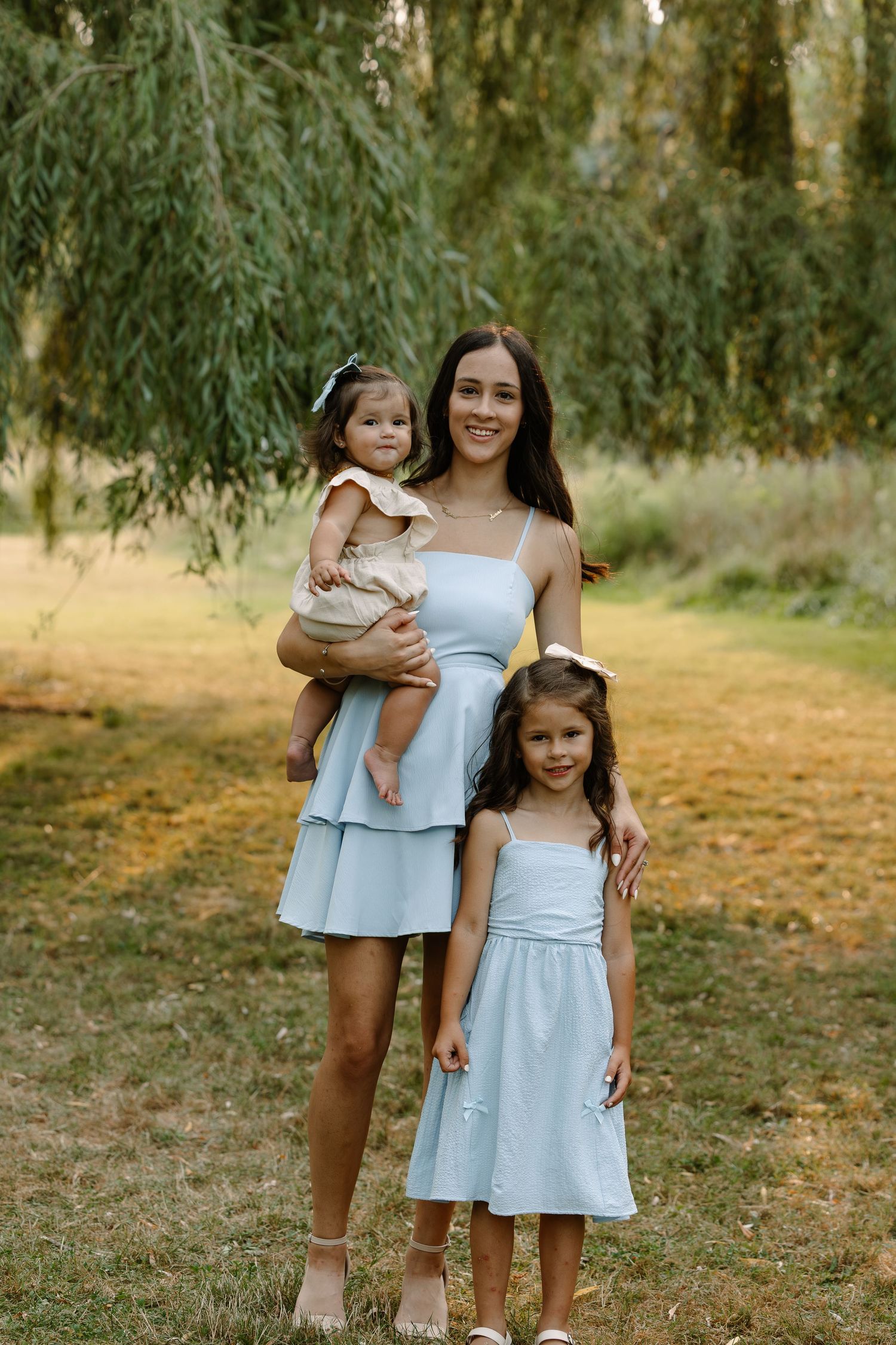 A mother in a light blue dress holds a toddler while her young daughter stands beside her outdoors.
