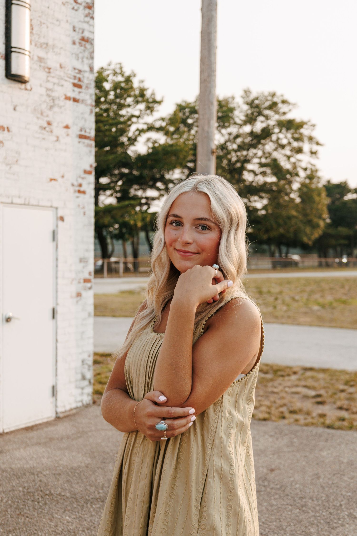 Young blonde woman in olive maxi dress posing outdoors near white brick building with turquoise ring.