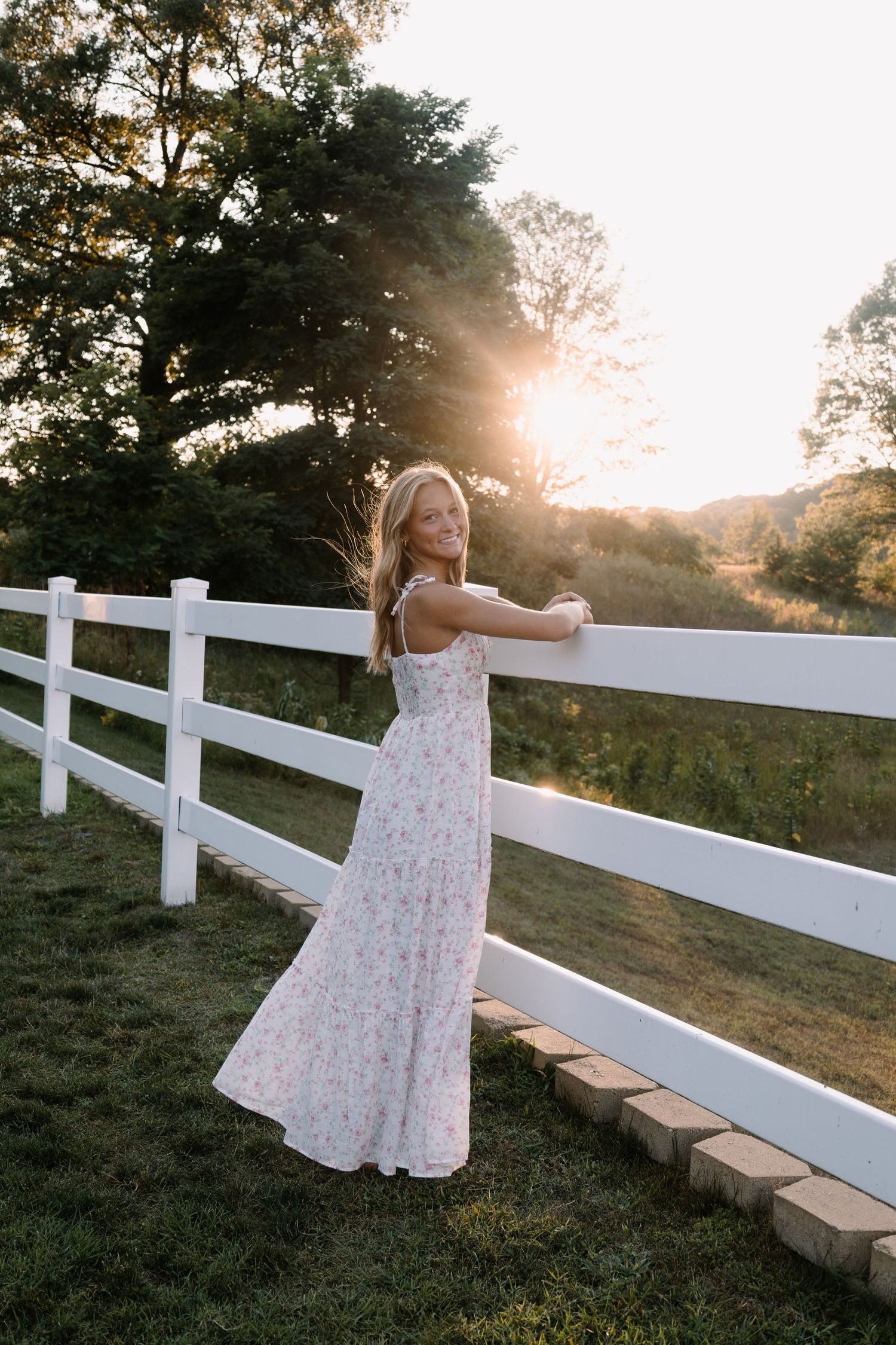 Blonde woman in a floral maxi dress leaning on a white fence at sunset with golden light streaming through the trees.