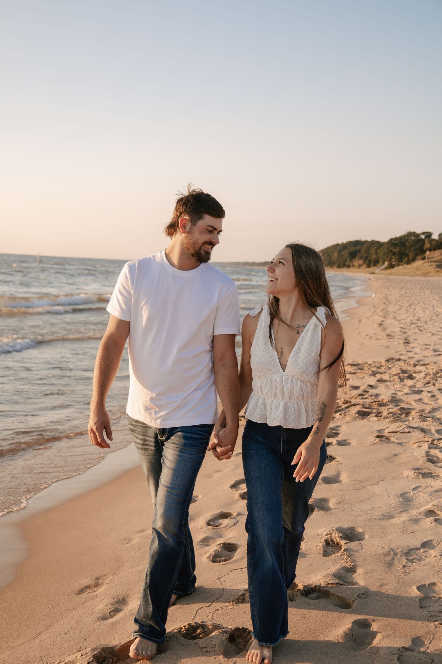 Couple holding hands and smiling at each other while walking barefoot on a sandy beach at sunset.