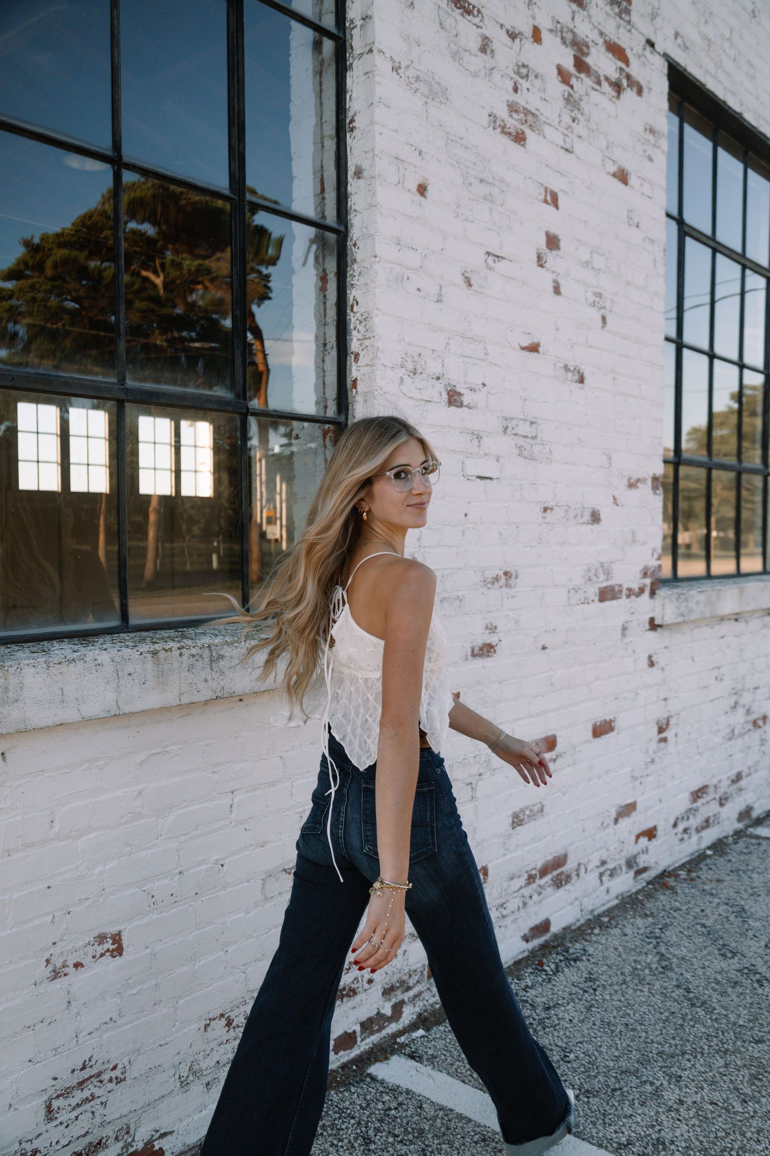 Blonde woman in white tank top and dark flare jeans walking beside a white brick building with industrial windows.