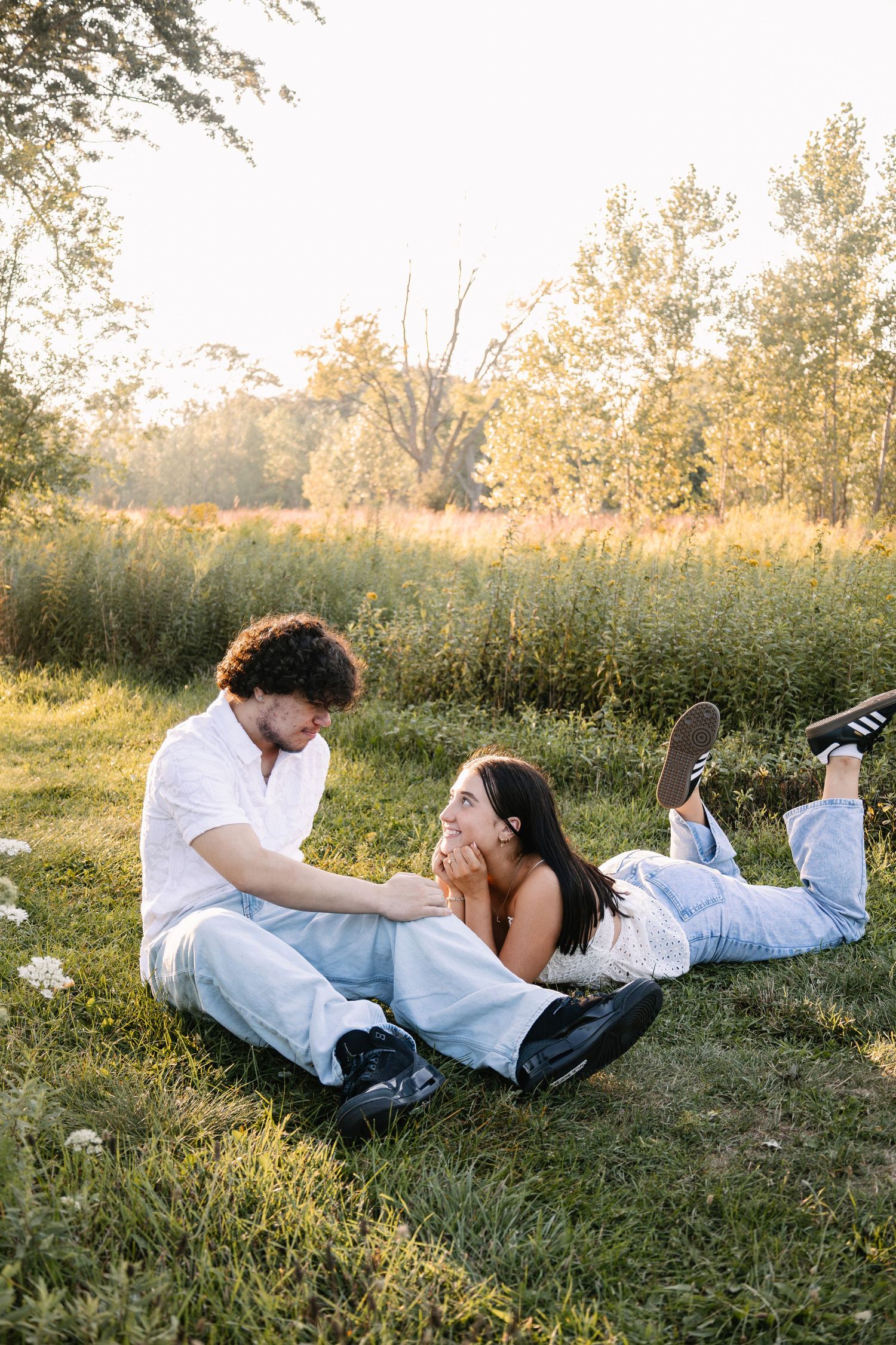 Couple relaxing on grass at golden hour, woman lying down laughing, man sitting beside her in a lush green park.
