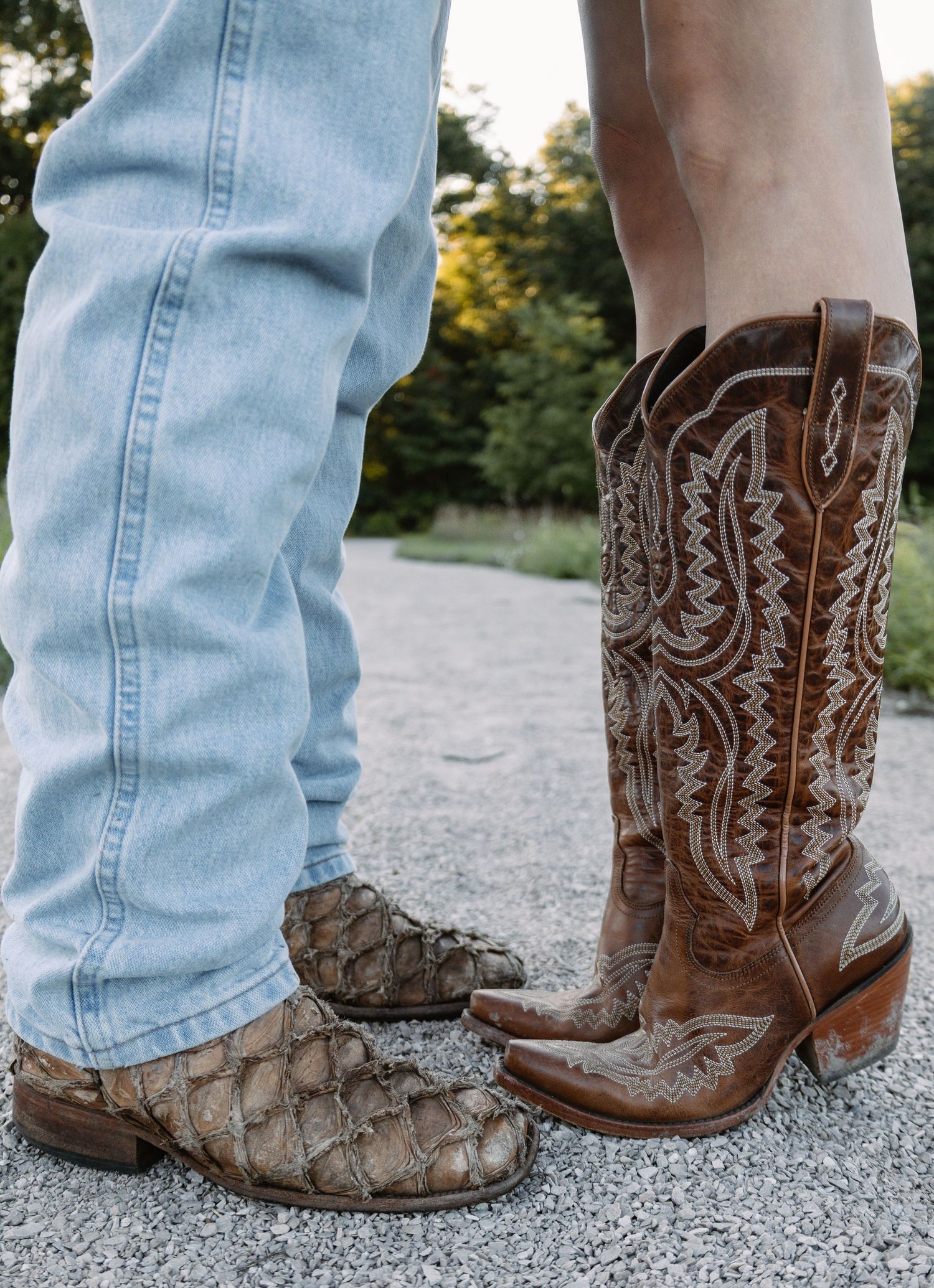 Two people wearing cowboy boots on a gravel path, one in snakeskin and one in brown embroidered leather.