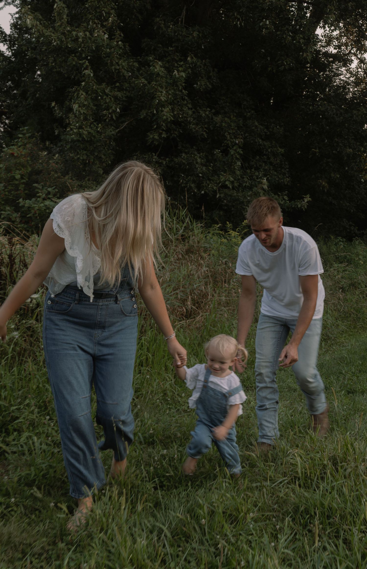 A young family with a toddler walks through a lush green meadow, parents holding the child's hands.
