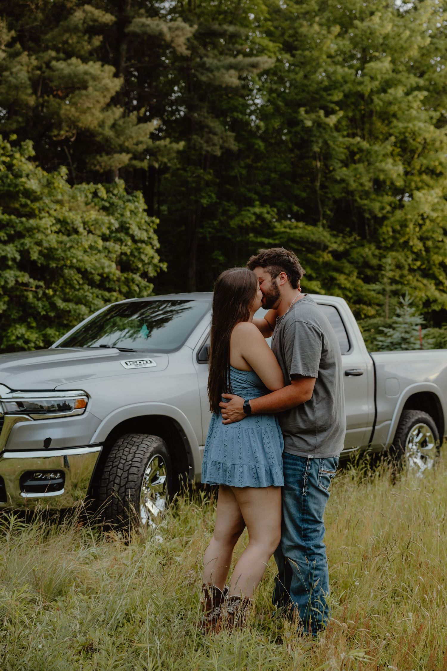 Romantic couple embracing and kissing in front of a silver Ram pickup truck in a forest meadow.