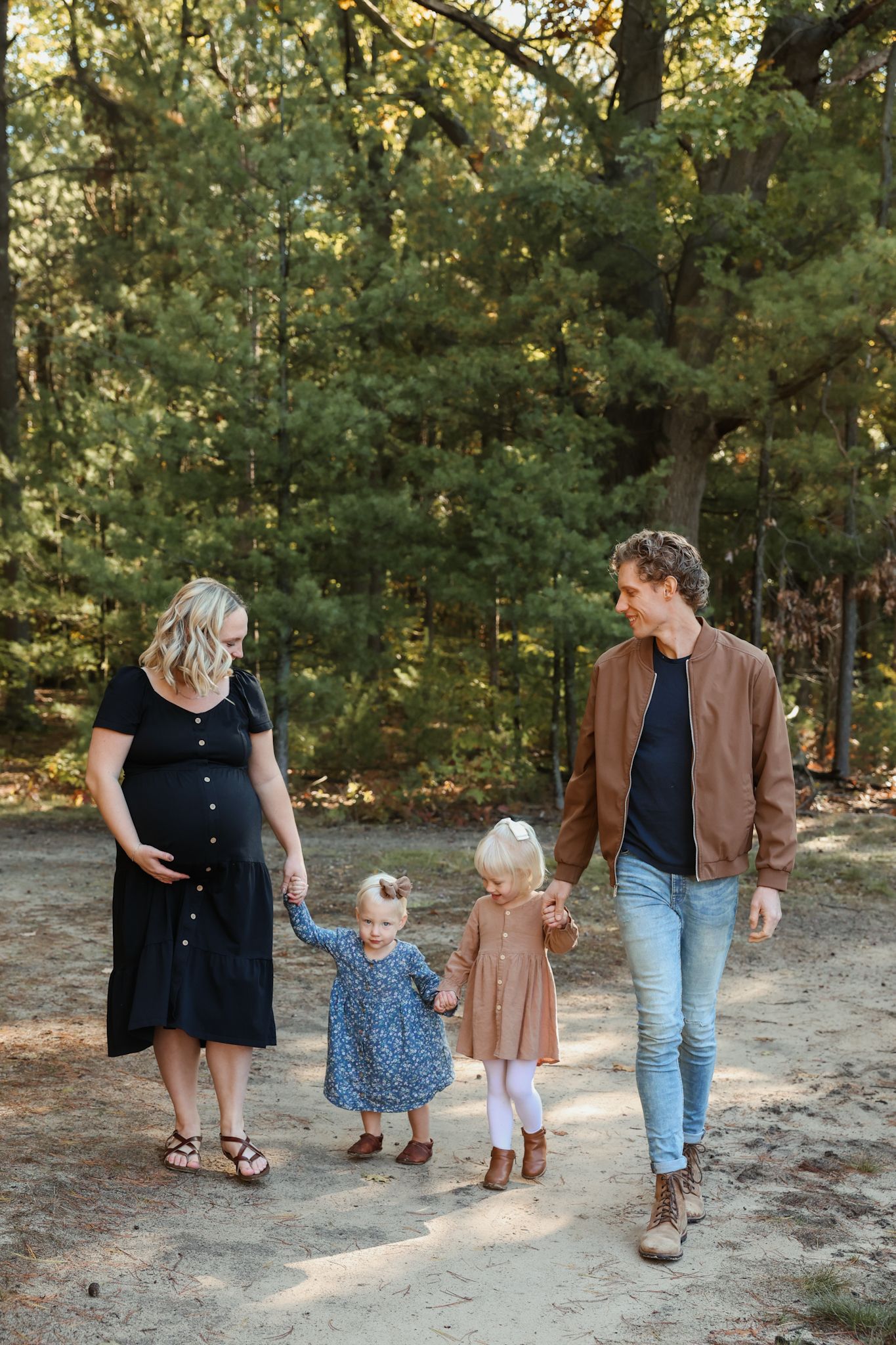 Pregnant mother and father walking with two young daughters hand in hand through a forest path.
