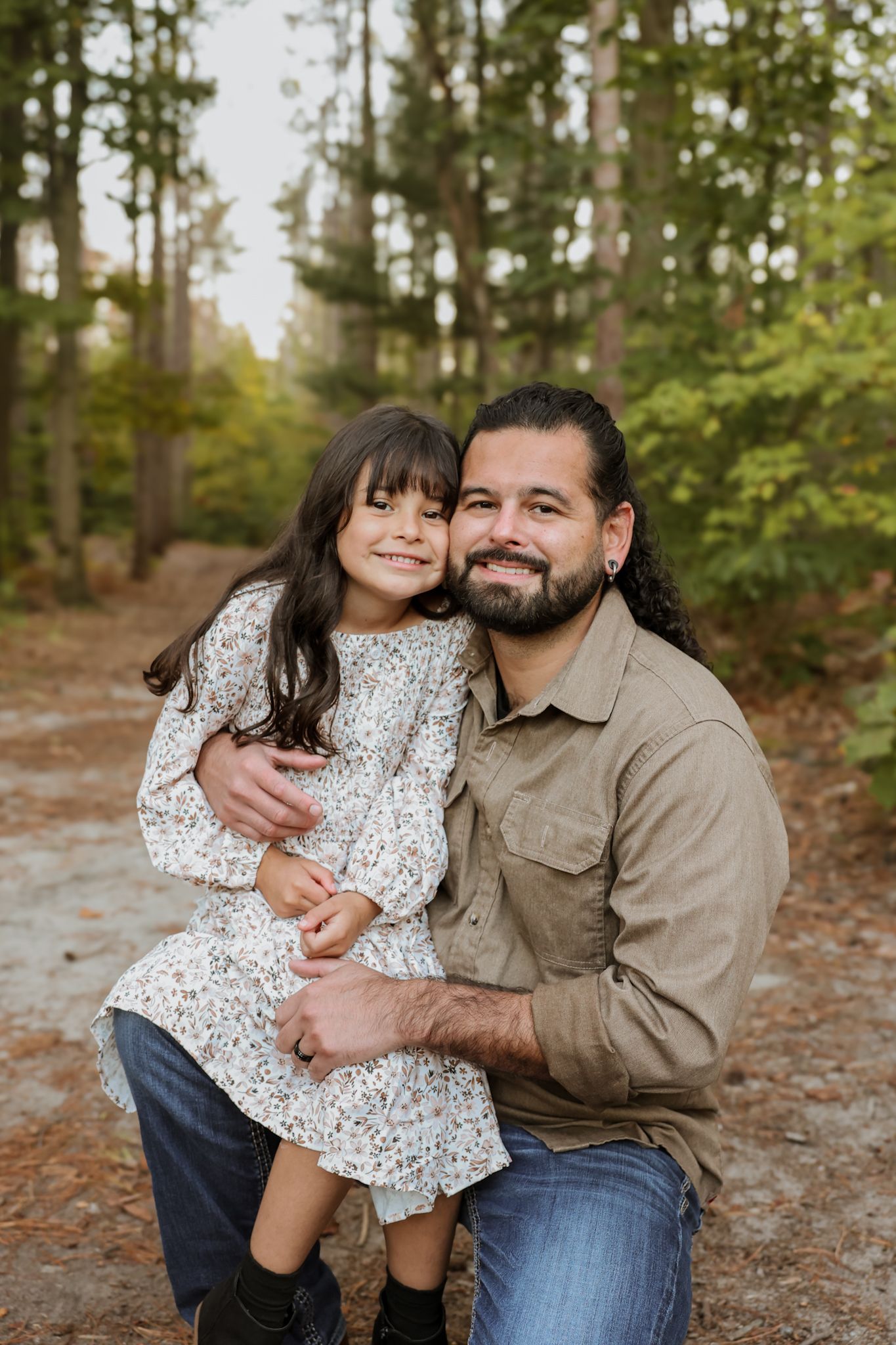 Smiling bearded father kneeling and hugging his young daughter on a forest path.