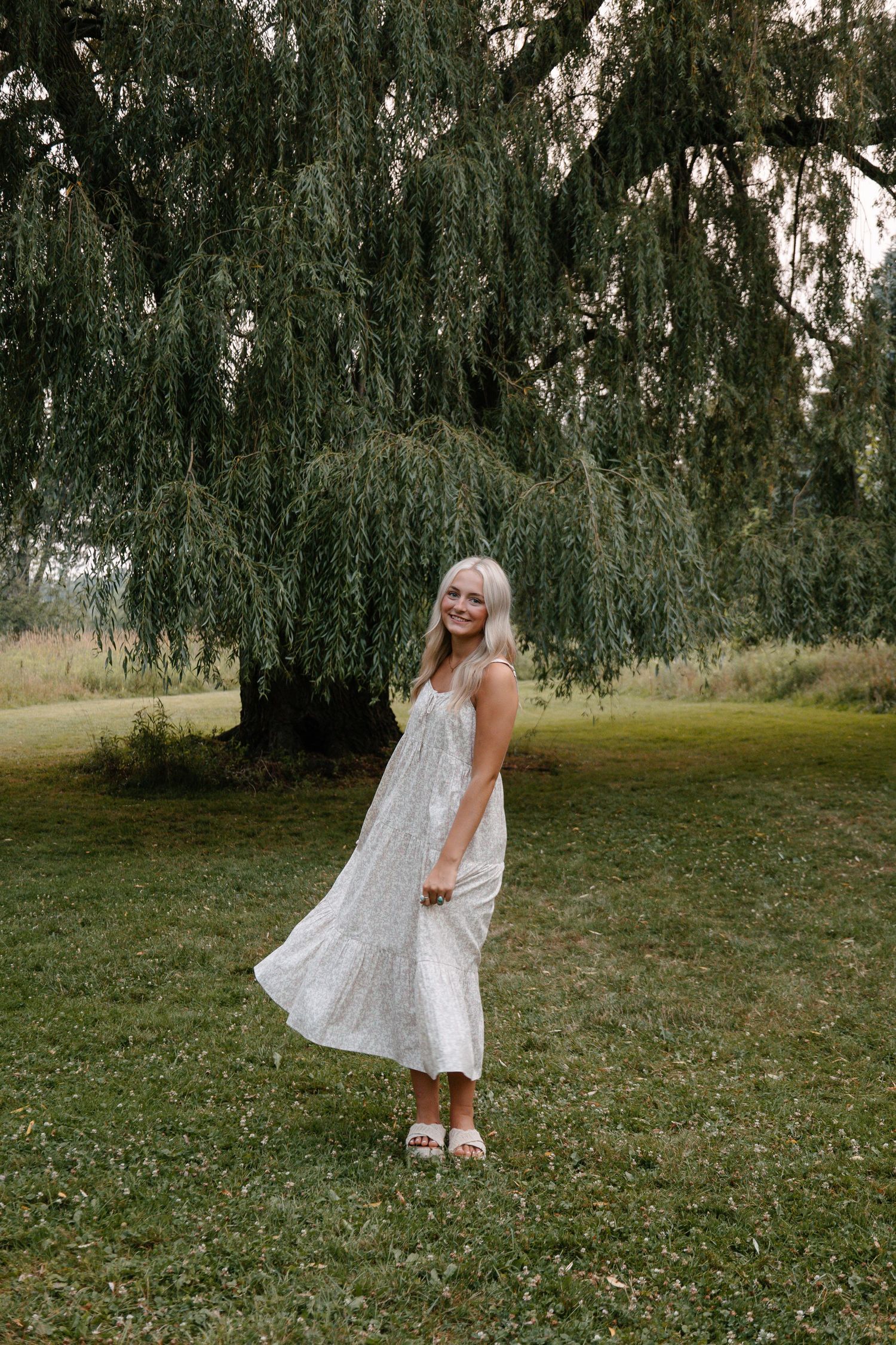 Young blonde woman in white floral midi dress twirling under a weeping willow tree on green grass.