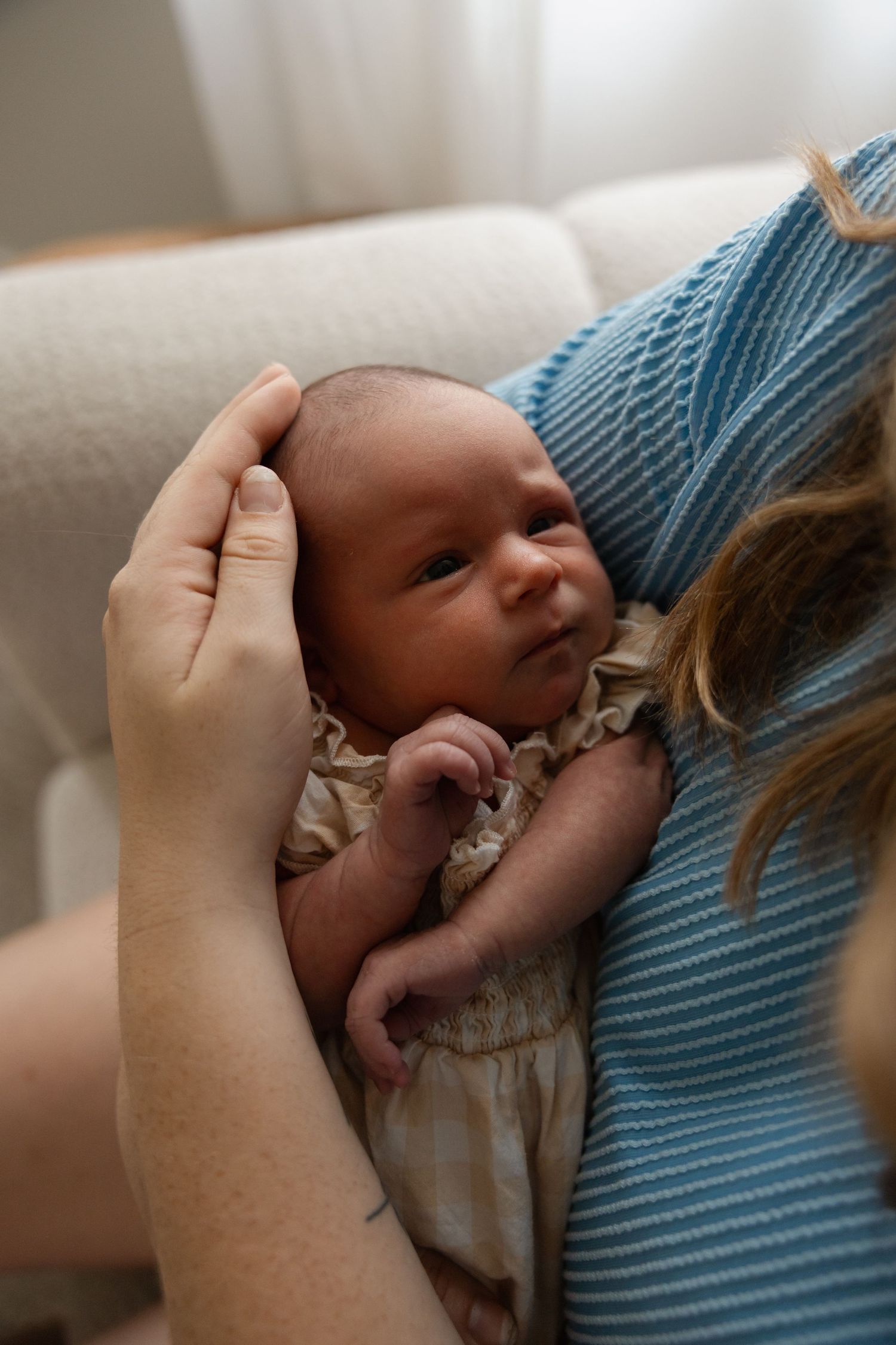 A newborn baby cradled in a mother's arms, wearing a cream outfit, with a gentle hand supporting its head.