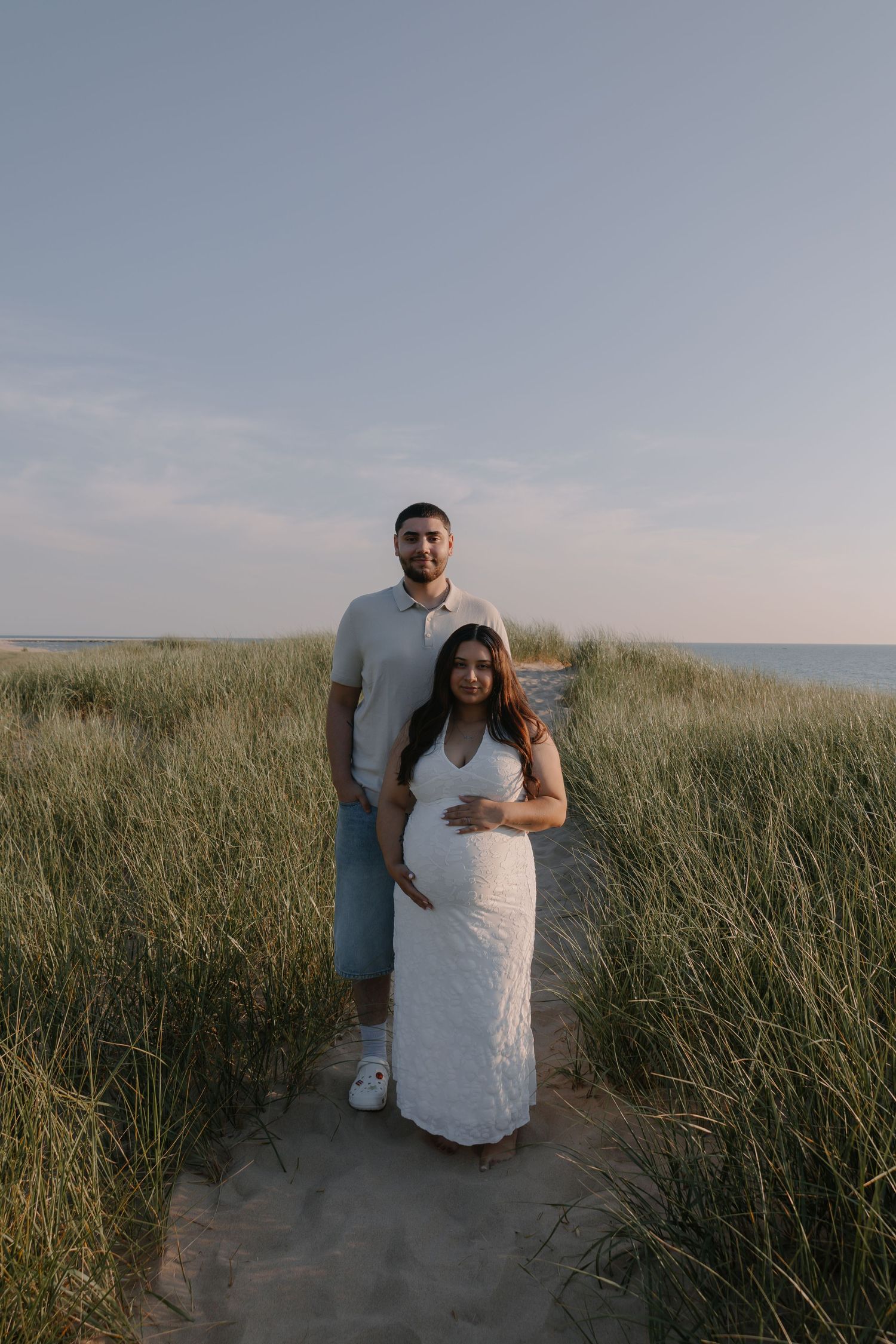 Couple poses on sandy beach path, pregnant woman in white lace dress, man in white shirt, ocean backdrop.