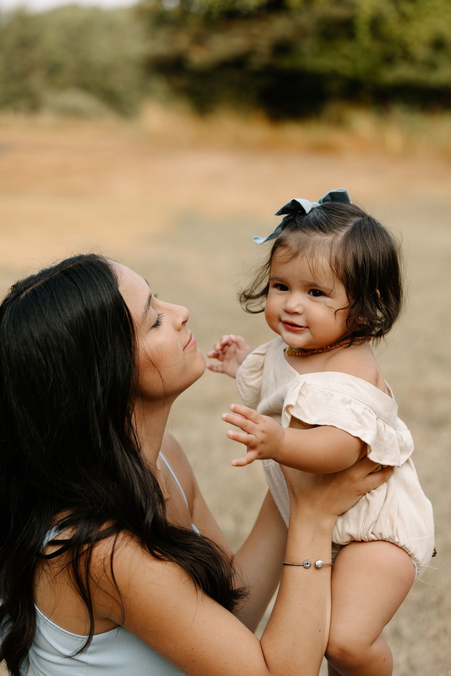 A mother with dark hair holds her smiling baby girl wearing a bow outdoors in a golden field.
