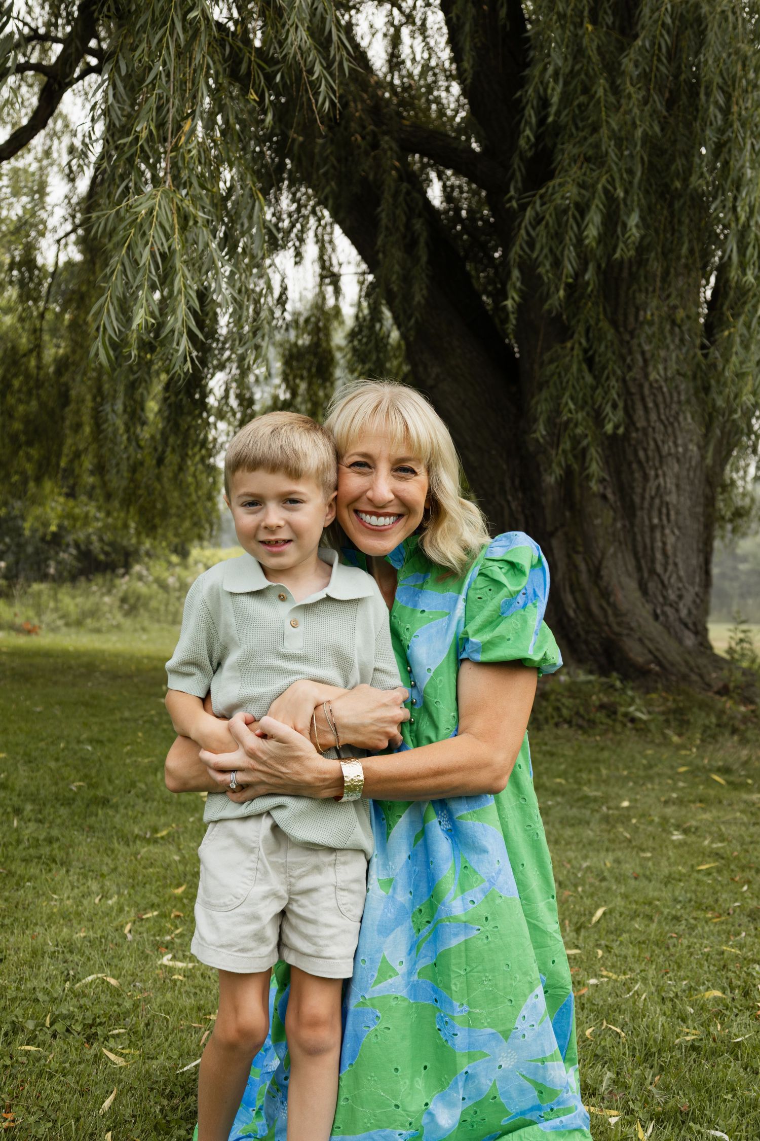 A smiling woman in a green dress hugs a young blonde boy outdoors near a large willow tree on a grassy lawn.