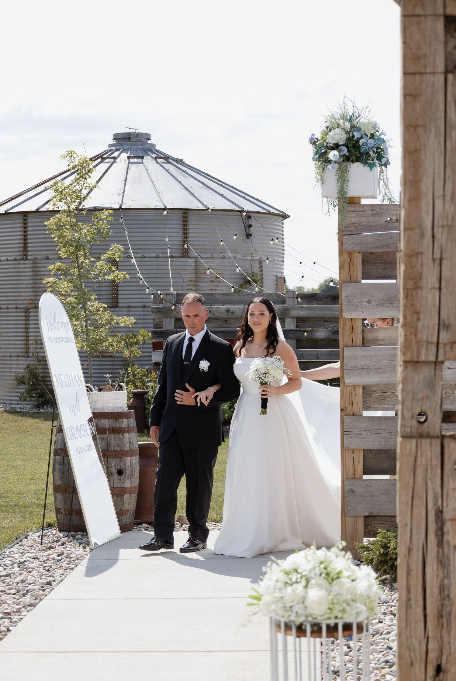Bride and groom walking down outdoor aisle at rustic barn wedding venue with grain silo backdrop.