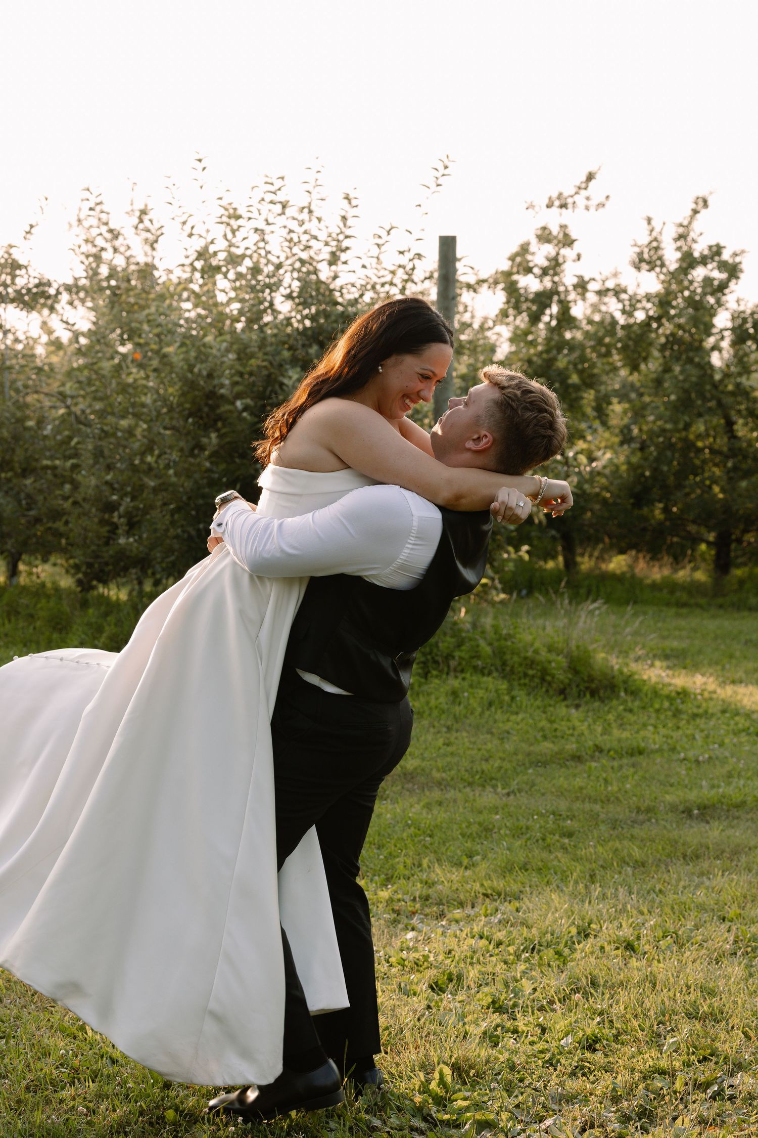 Groom lifts laughing bride in white strapless gown in a romantic embrace in an orchard.