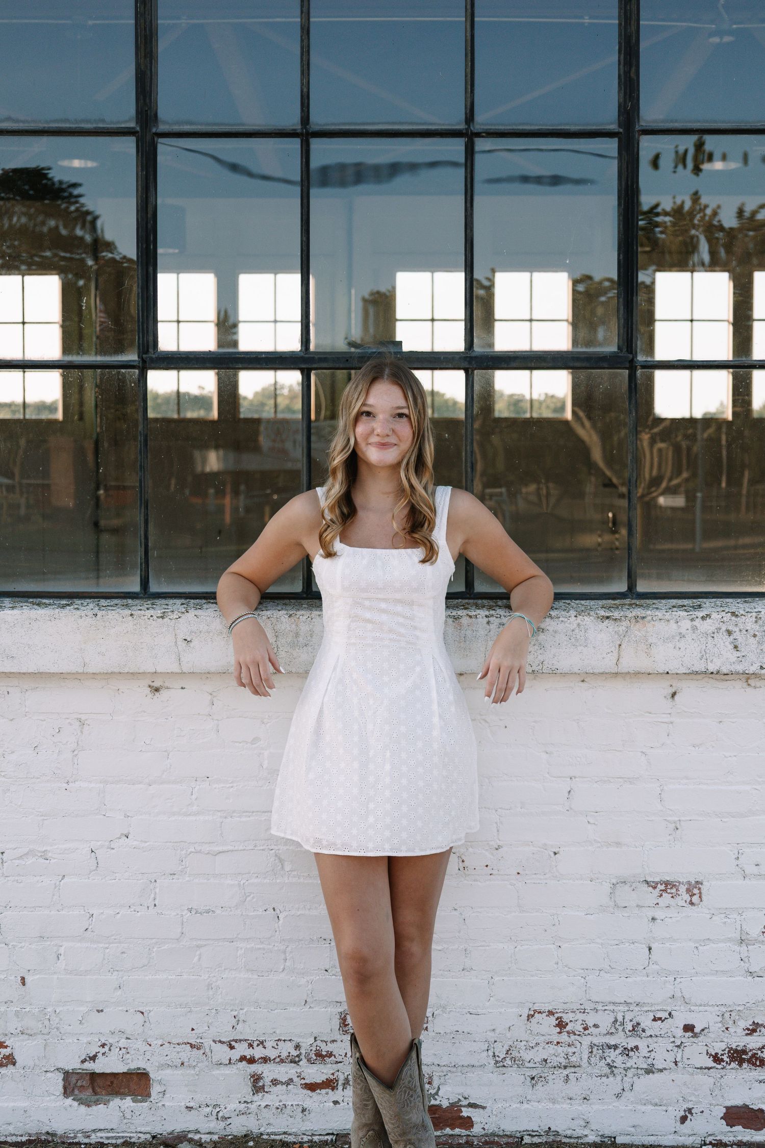 Young woman in white mini dress and cowboy boots posing by industrial window wall.