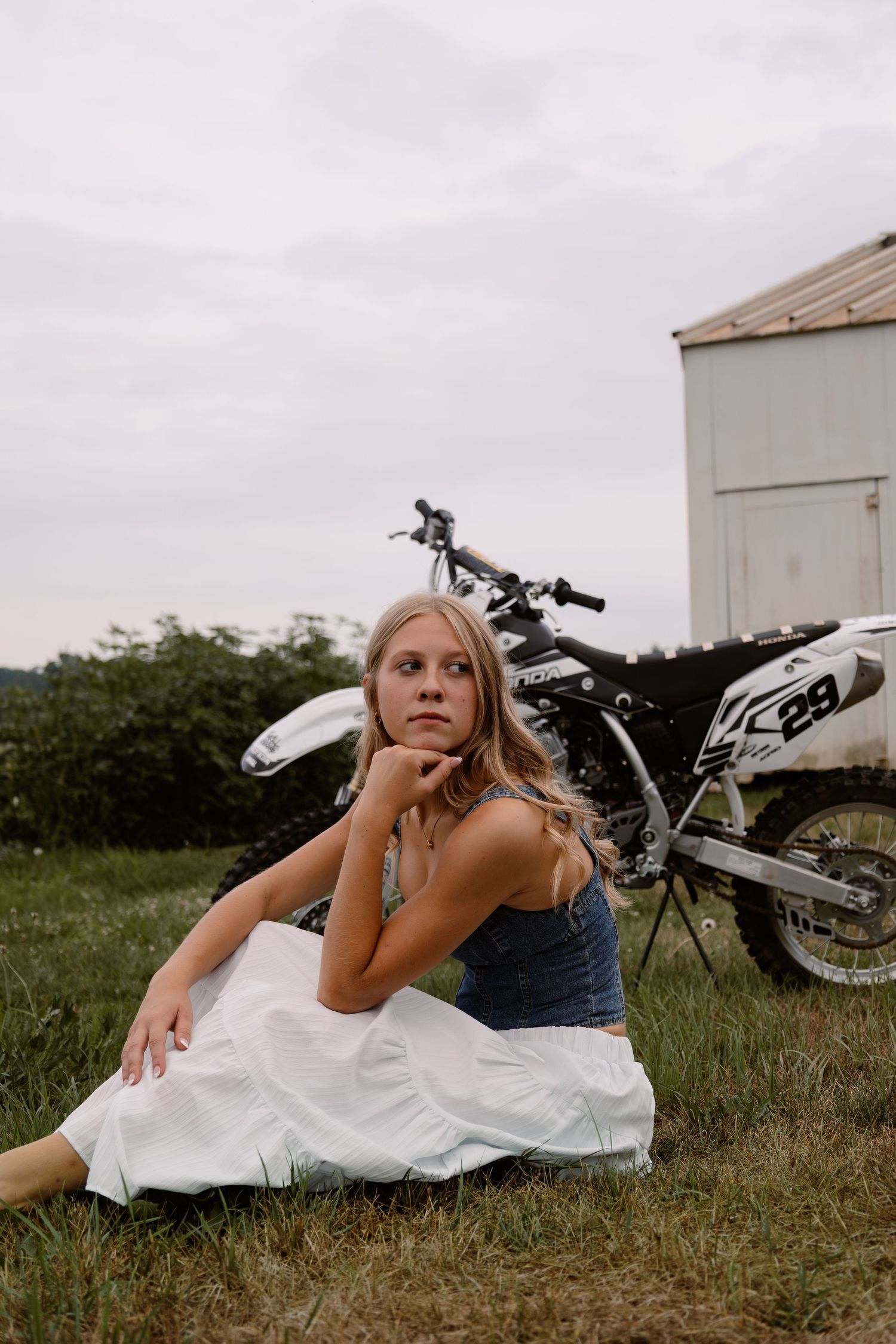 Young woman in white skirt and denim top sitting on grass beside a white dirt bike numbered 29.