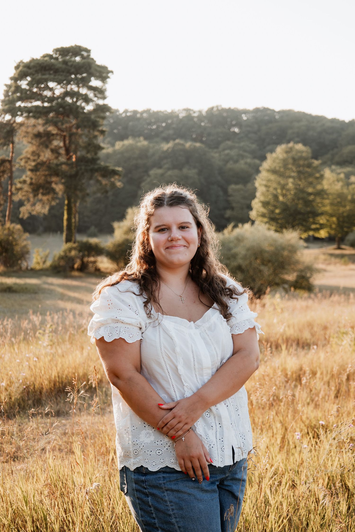 Young woman in white eyelet top and jeans smiling in a golden field with trees and hills behind her.
