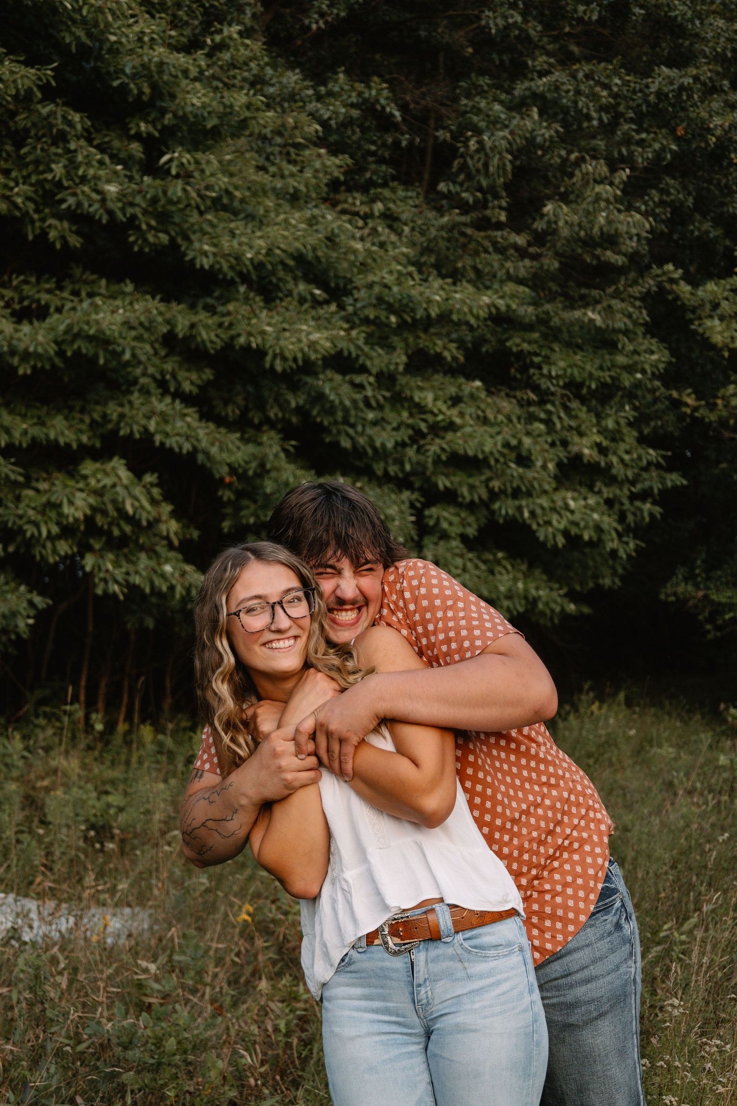A smiling couple embraces outdoors, man in polka dot shirt hugging woman in white top and jeans, with green trees behind.