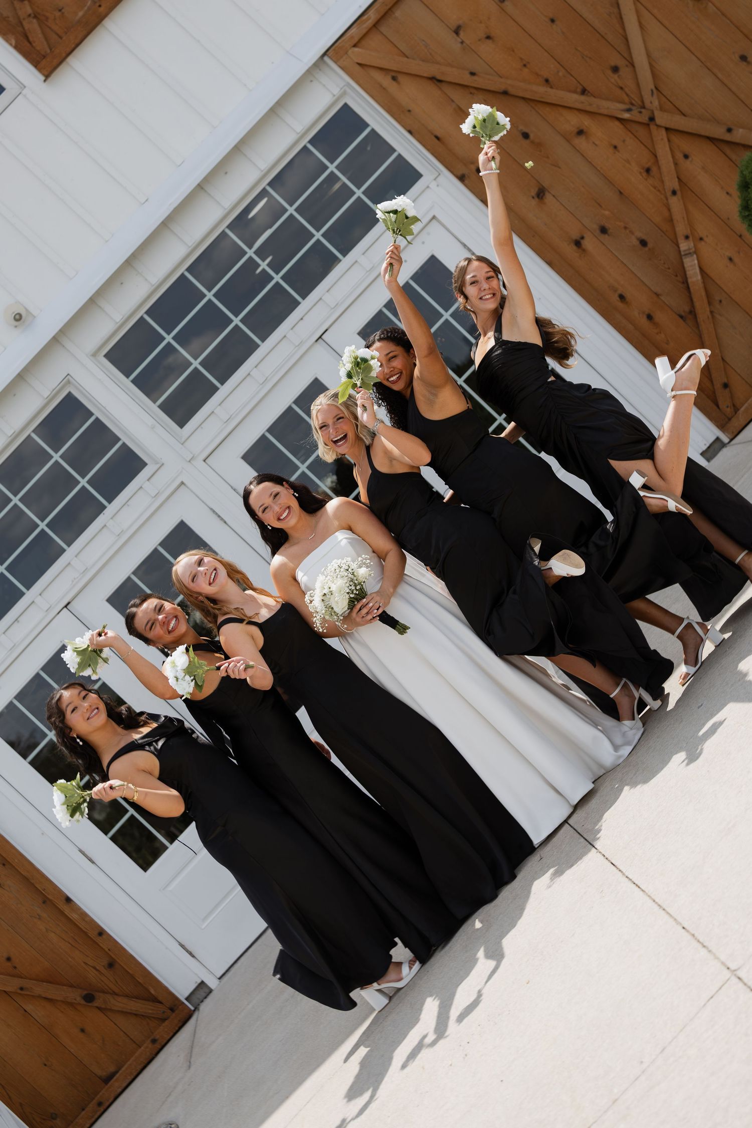 Bride in white gown with bridesmaids in black dresses jumping joyfully outside a white barn venue.