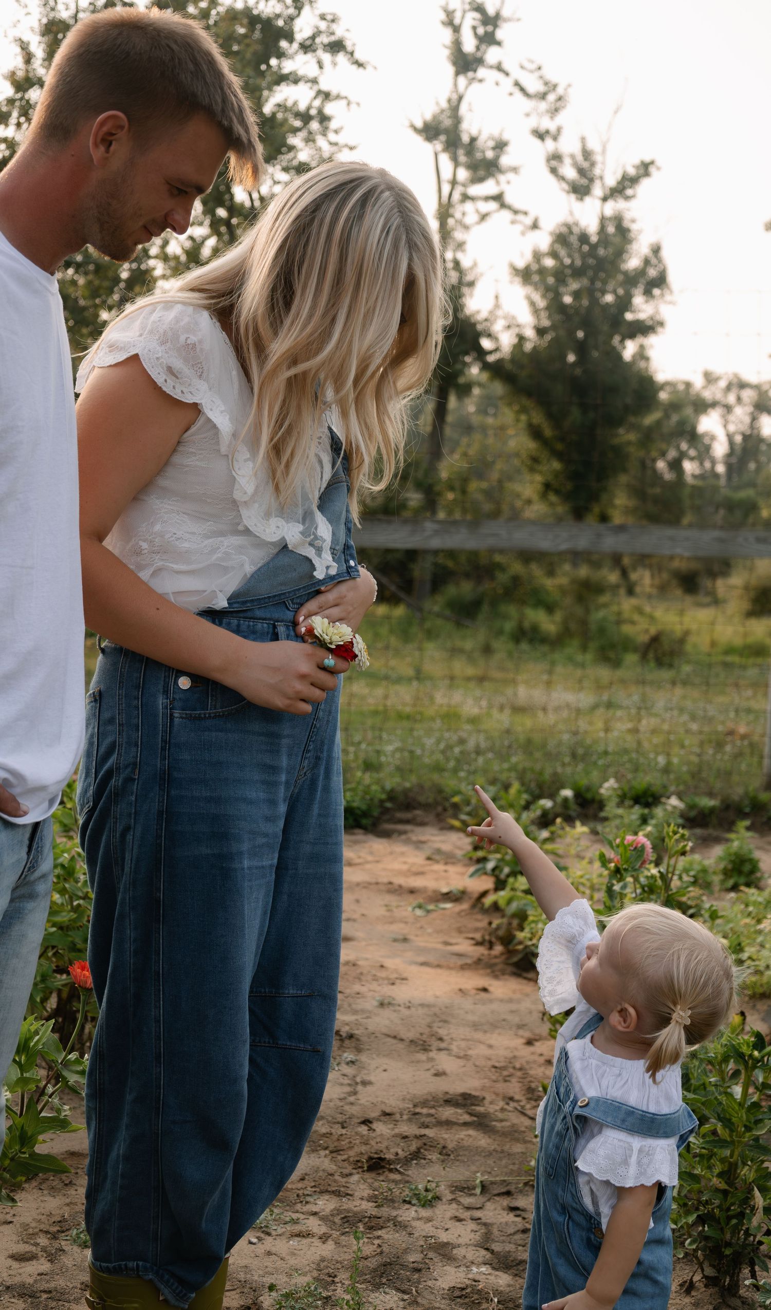 A family in denim overalls stands in a flower garden, with a toddler pointing up at her parents.