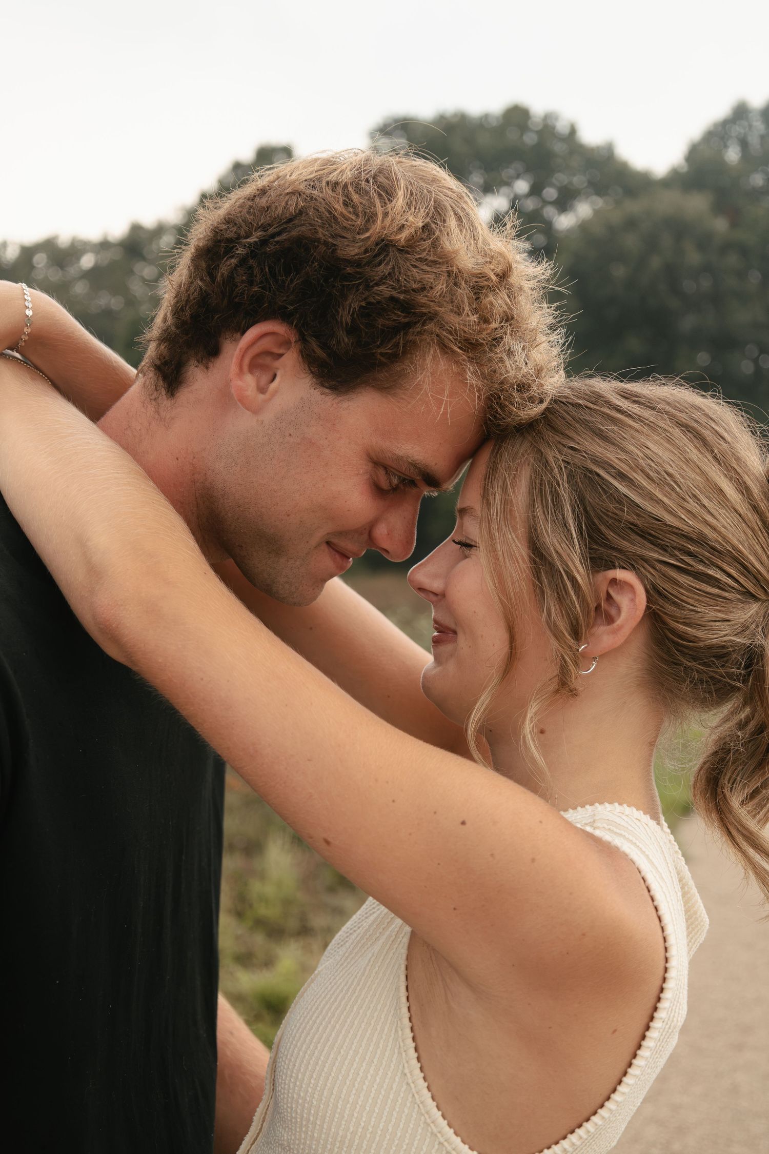 A young couple shares a romantic moment outdoors, foreheads touching and smiling, surrounded by lush green trees.