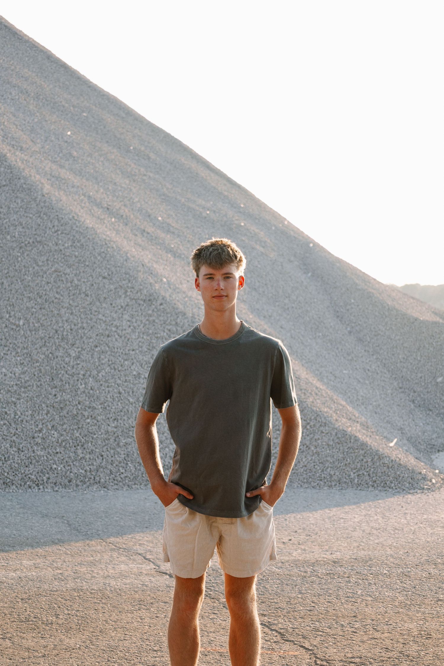 Young man in gray t-shirt and beige shorts stands confidently before a large gravel mound in natural light.