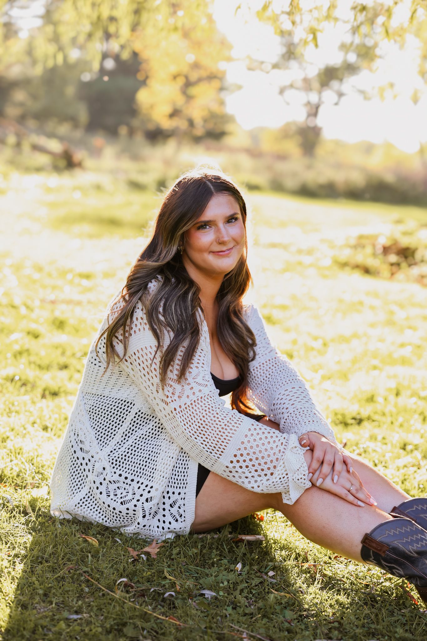 Young woman in white crochet cardigan sitting on grass in a sunny outdoor park setting.
