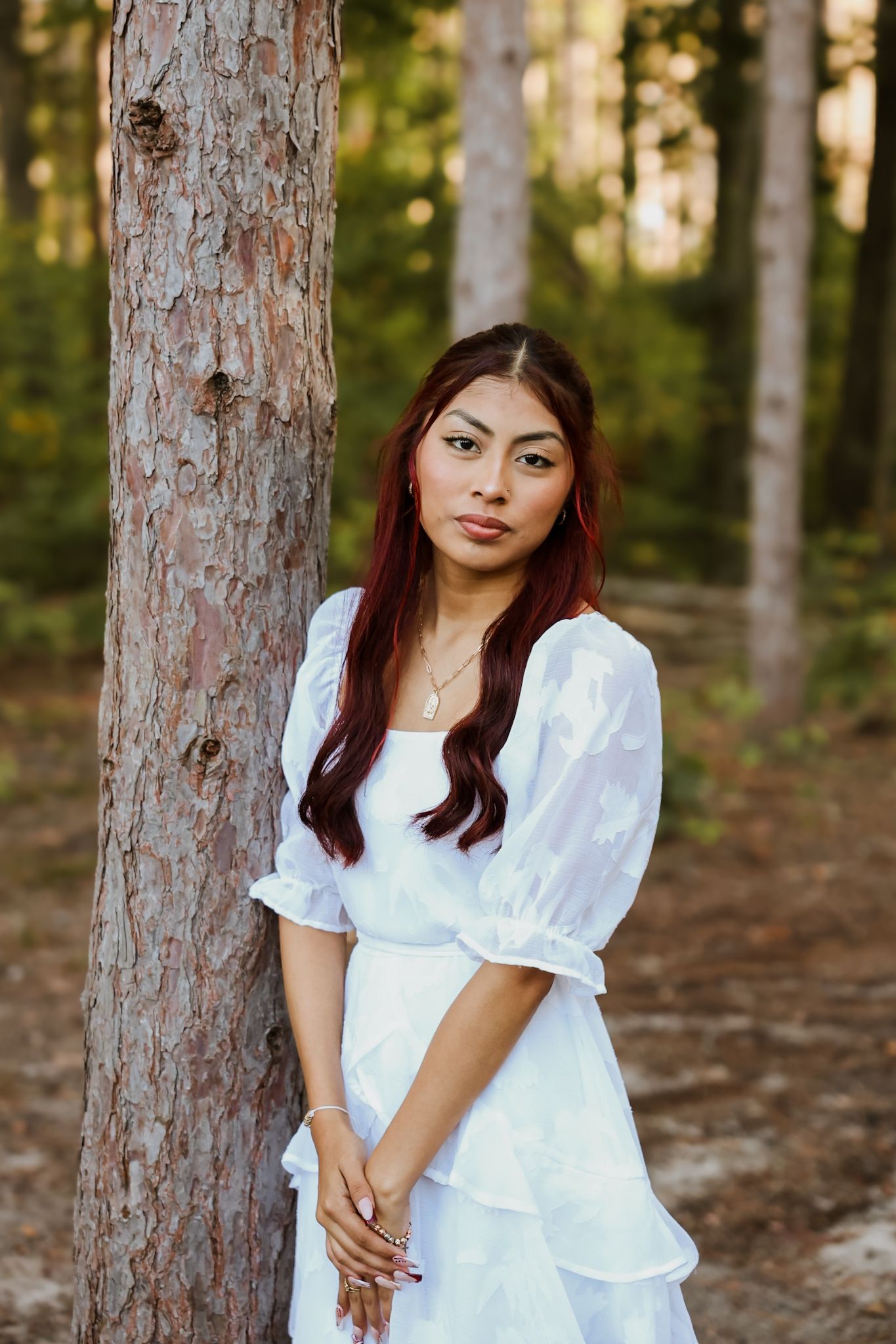Young woman in white dress leaning against pine tree in forest.