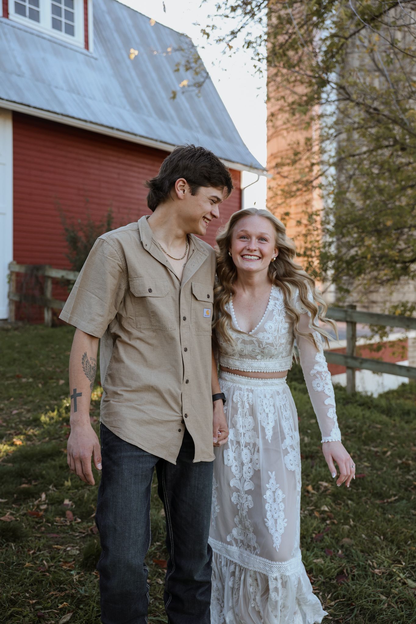 Happy engaged couple laughing and holding hands while walking near a red barn and silo.