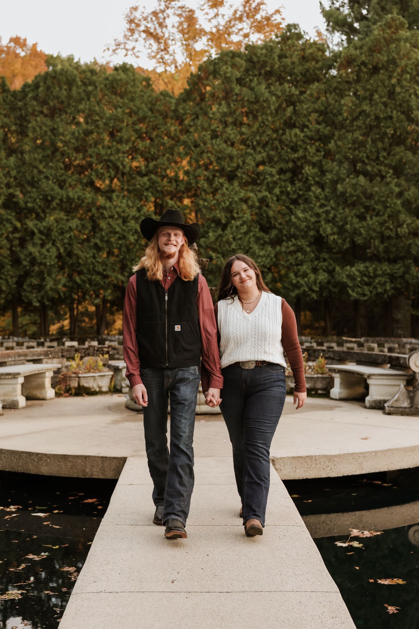 Couple holding hands walking on stone path by fountain in autumn park setting.