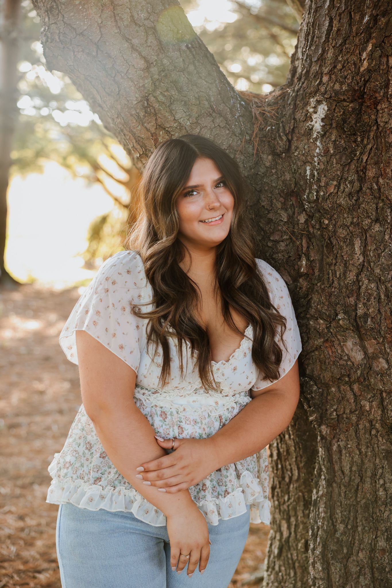 Smiling young woman in floral blouse leaning against a large tree trunk in warm sunlight.