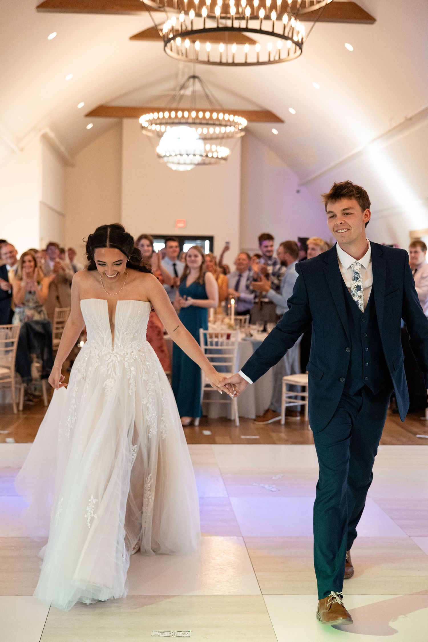 Bride and groom make grand entrance at elegant wedding reception hall with chandeliers and cheering guests.