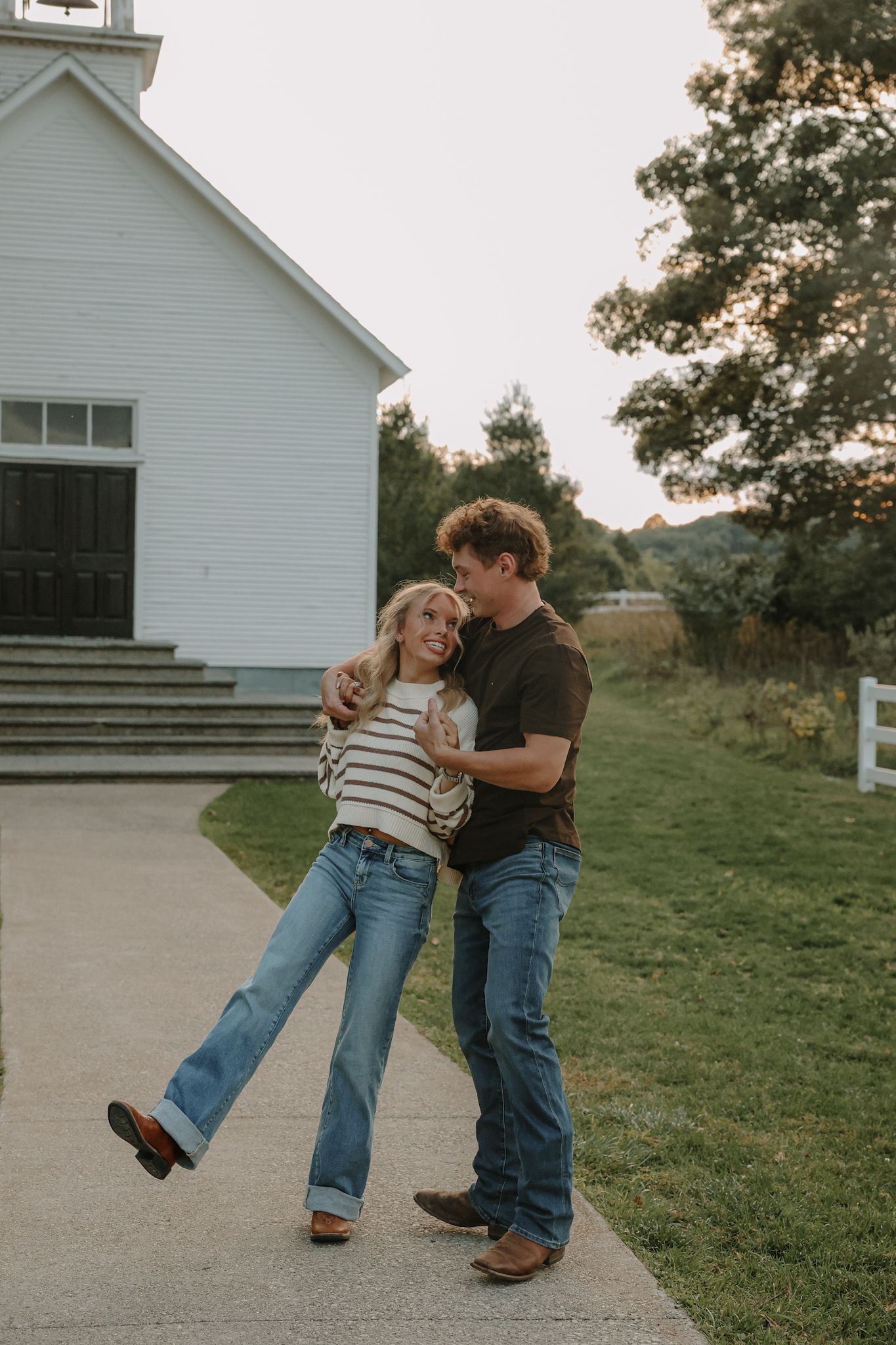 A joyful couple embraces and laughs together on a pathway beside a white country church at sunset.