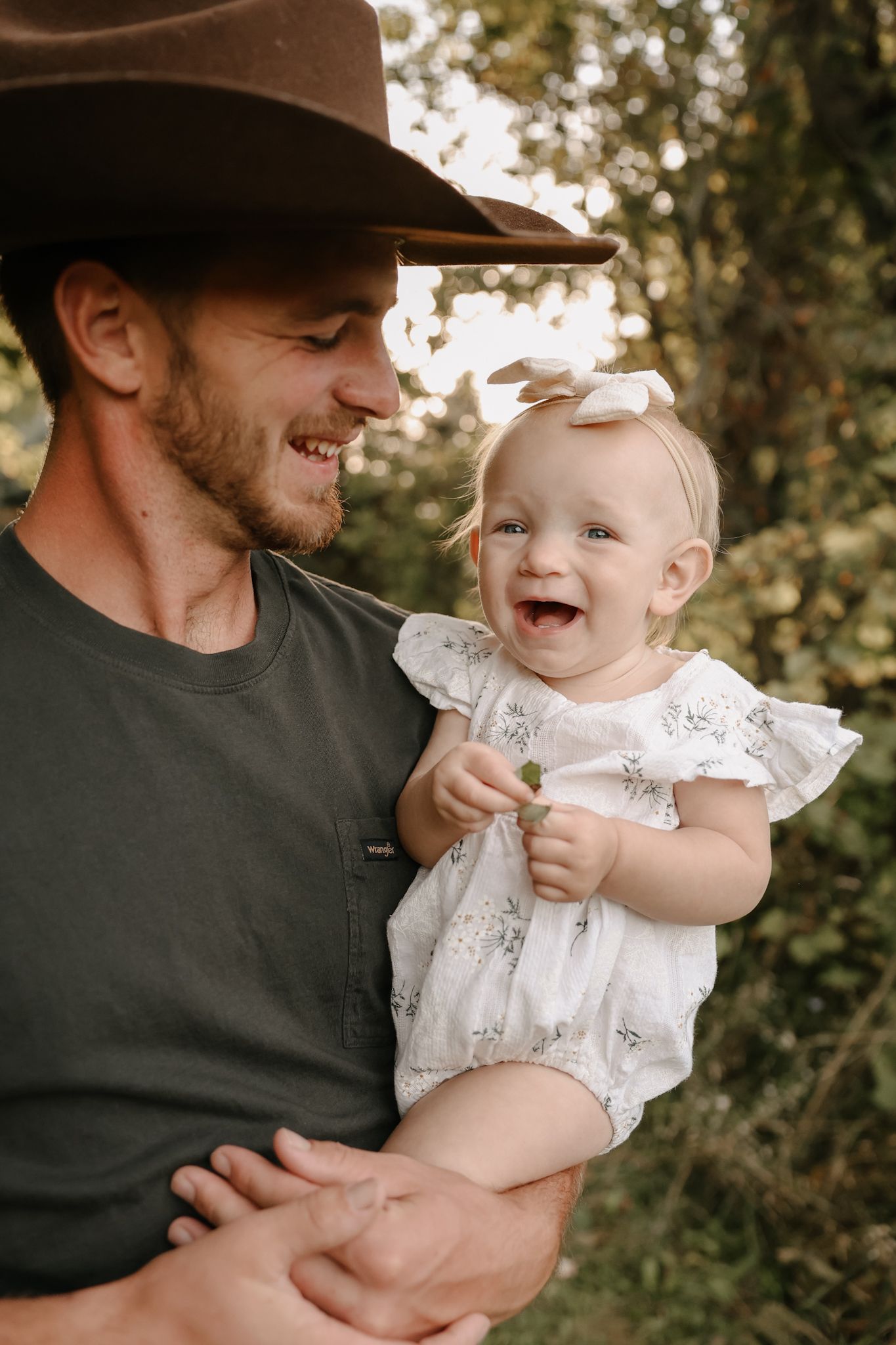Smiling dad in brown cowboy hat holding laughing baby girl in white romper in sunny outdoor setting.