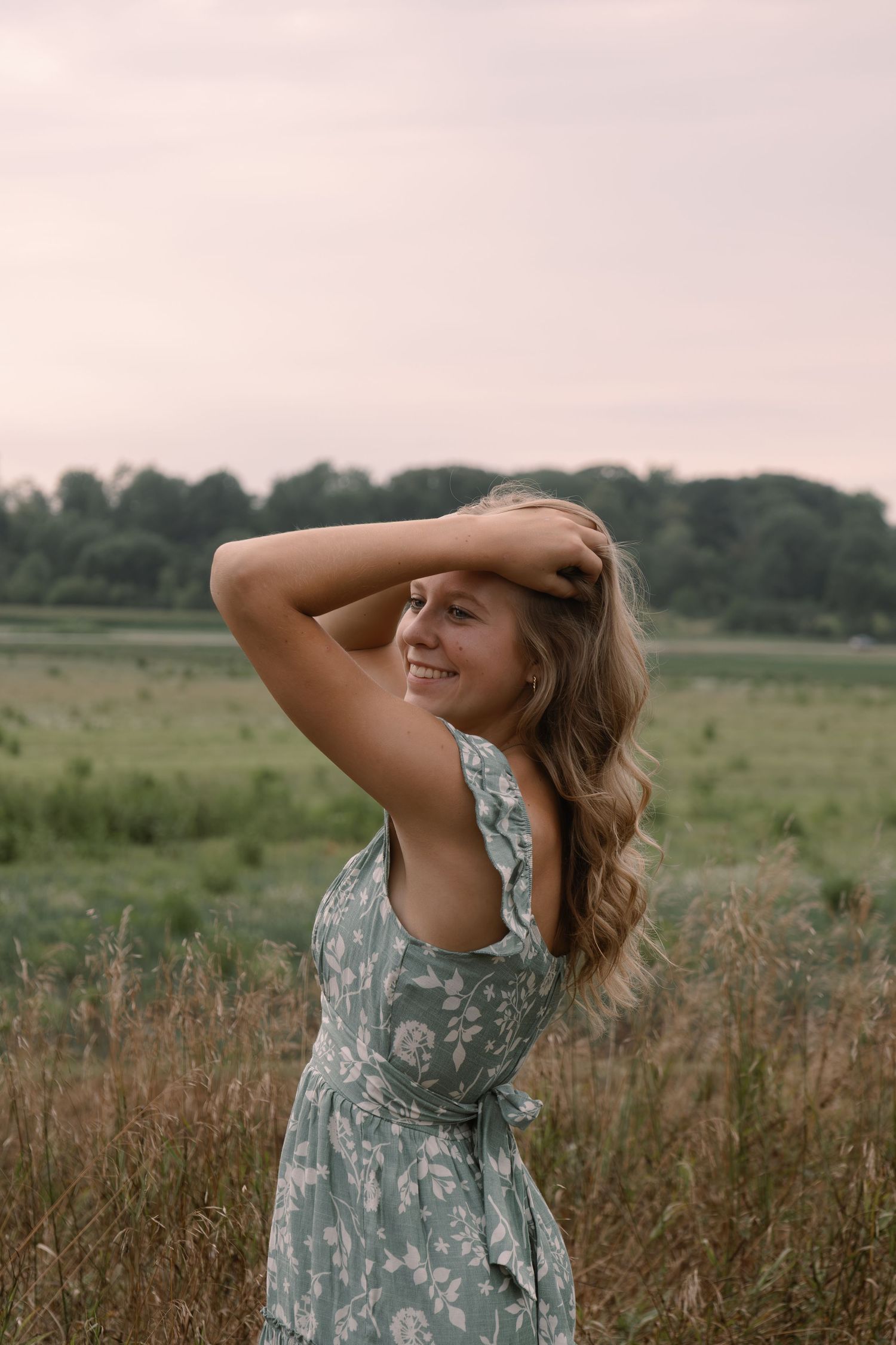 Smiling young woman in floral dress posing in a field at dusk, hand in hair.