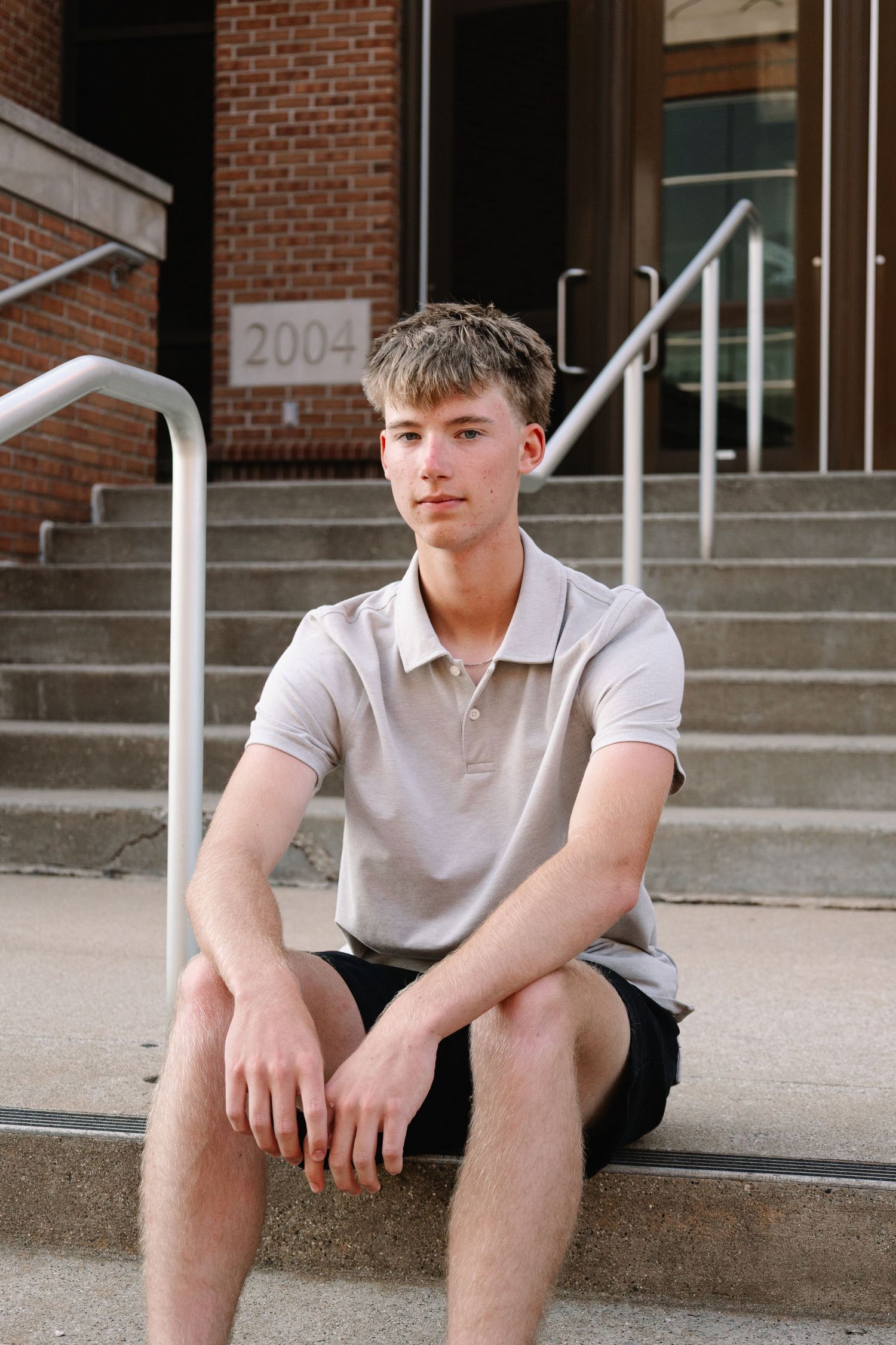 Young man in gray polo shirt and black shorts sitting on concrete steps outside a brick building.
