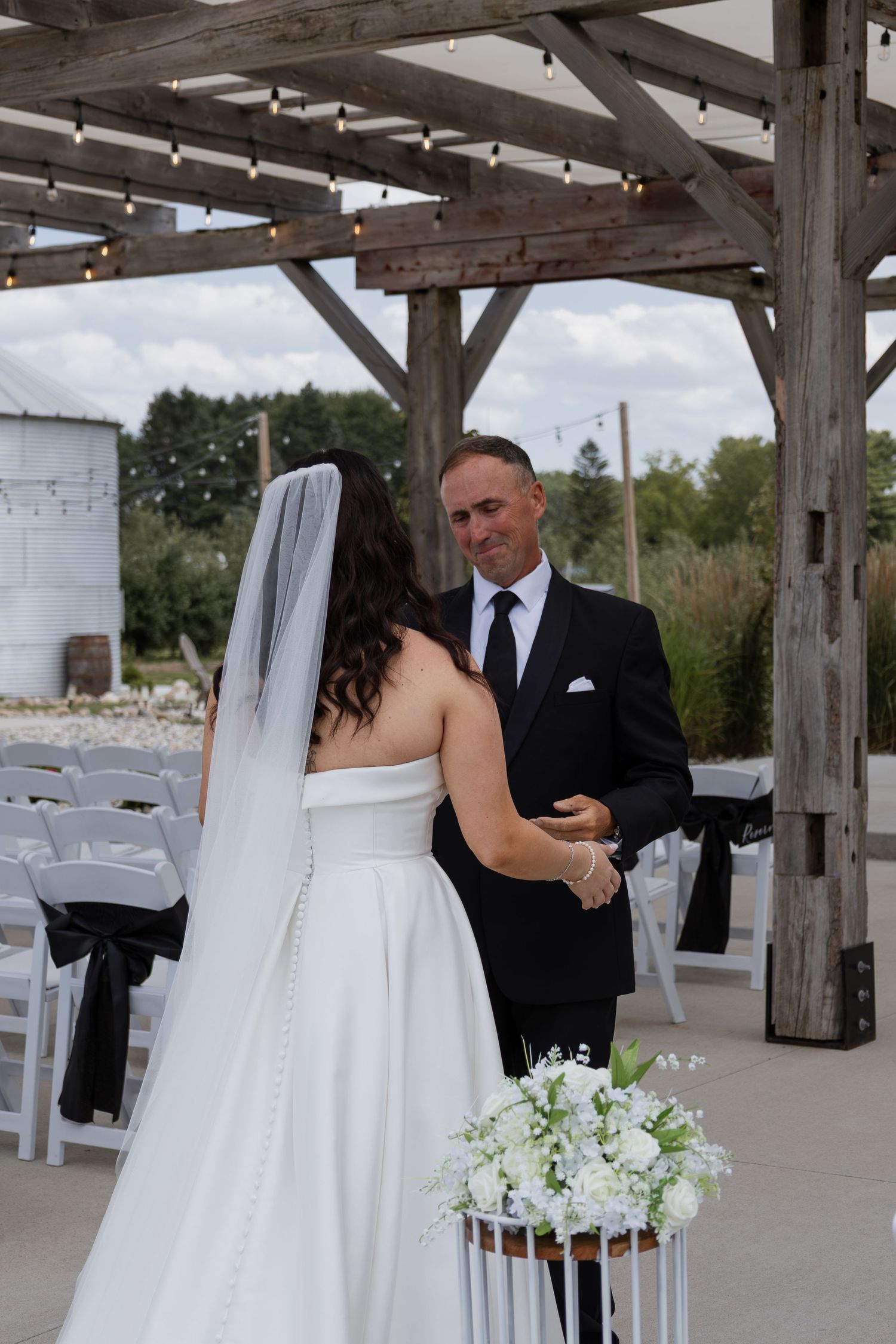 Bride and groom exchange vows under rustic wooden pergola at outdoor wedding ceremony with white chairs.