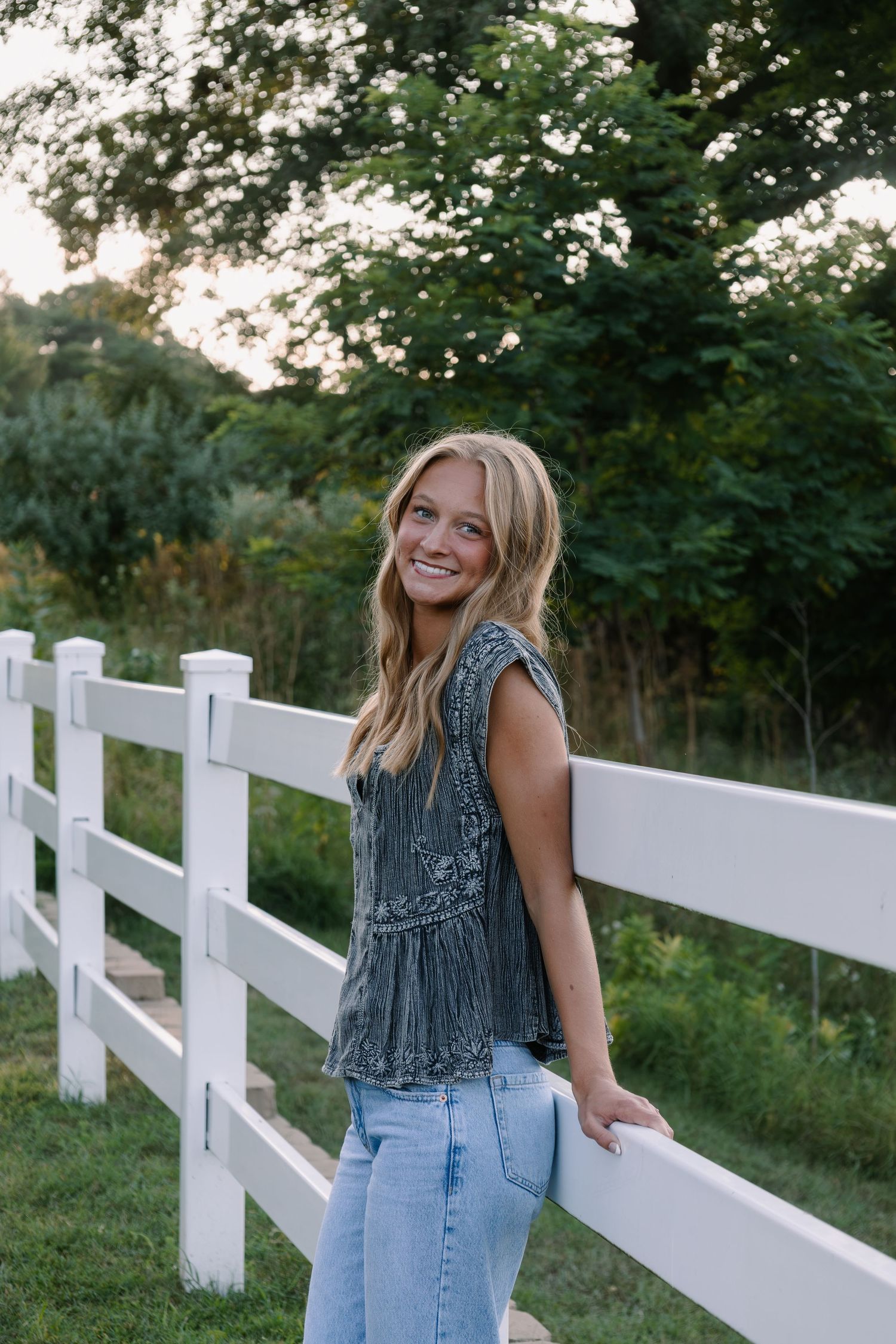 Smiling blonde teen girl in gray top and jeans leaning on white fence with green trees in background.