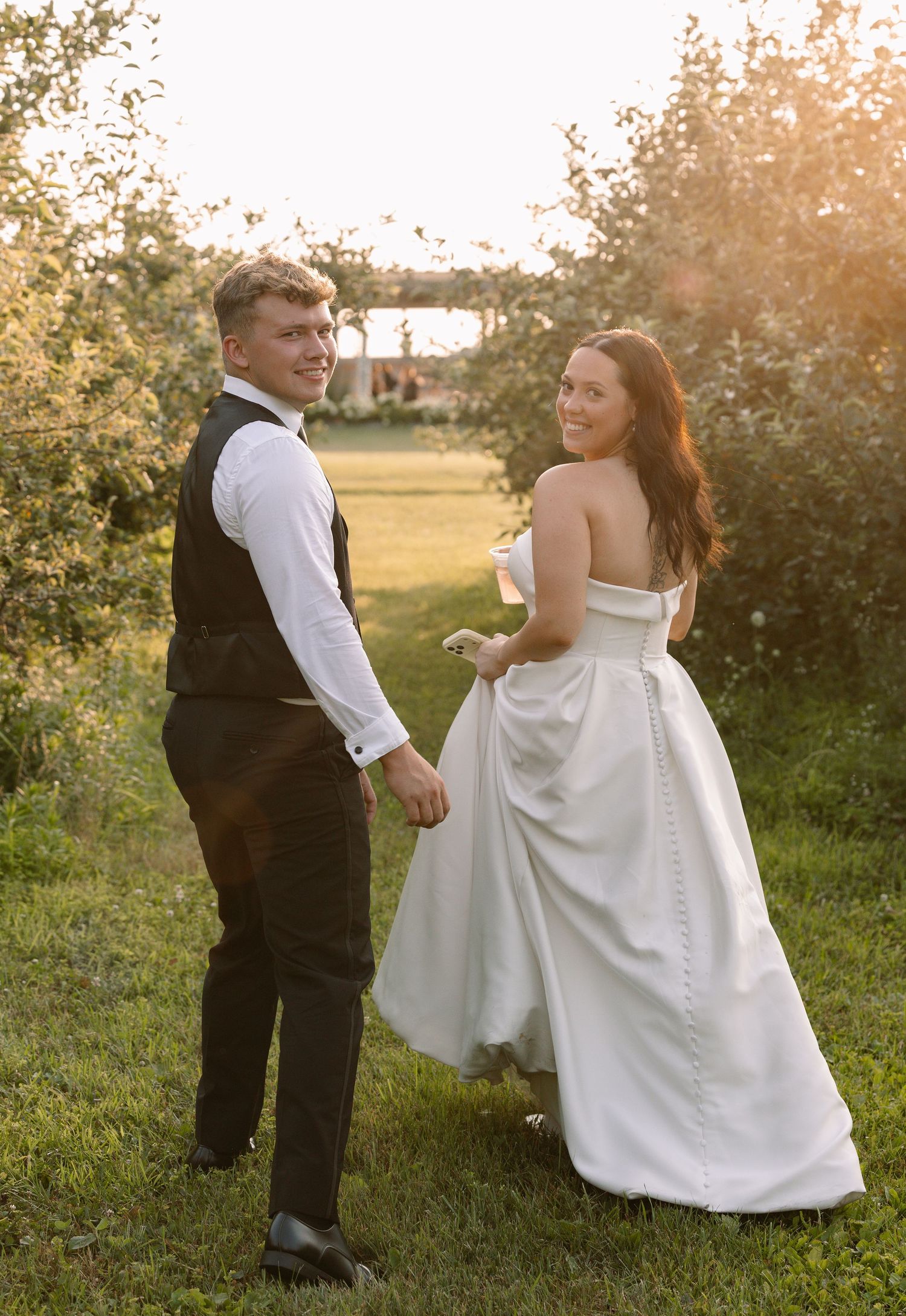 Bride and groom holding hands at golden hour in a lush green orchard, bride in strapless white gown.