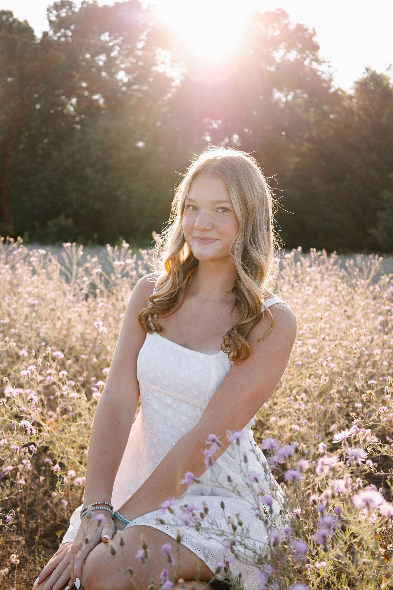 Young woman in white dress sitting in wildflower field at golden hour sunset.