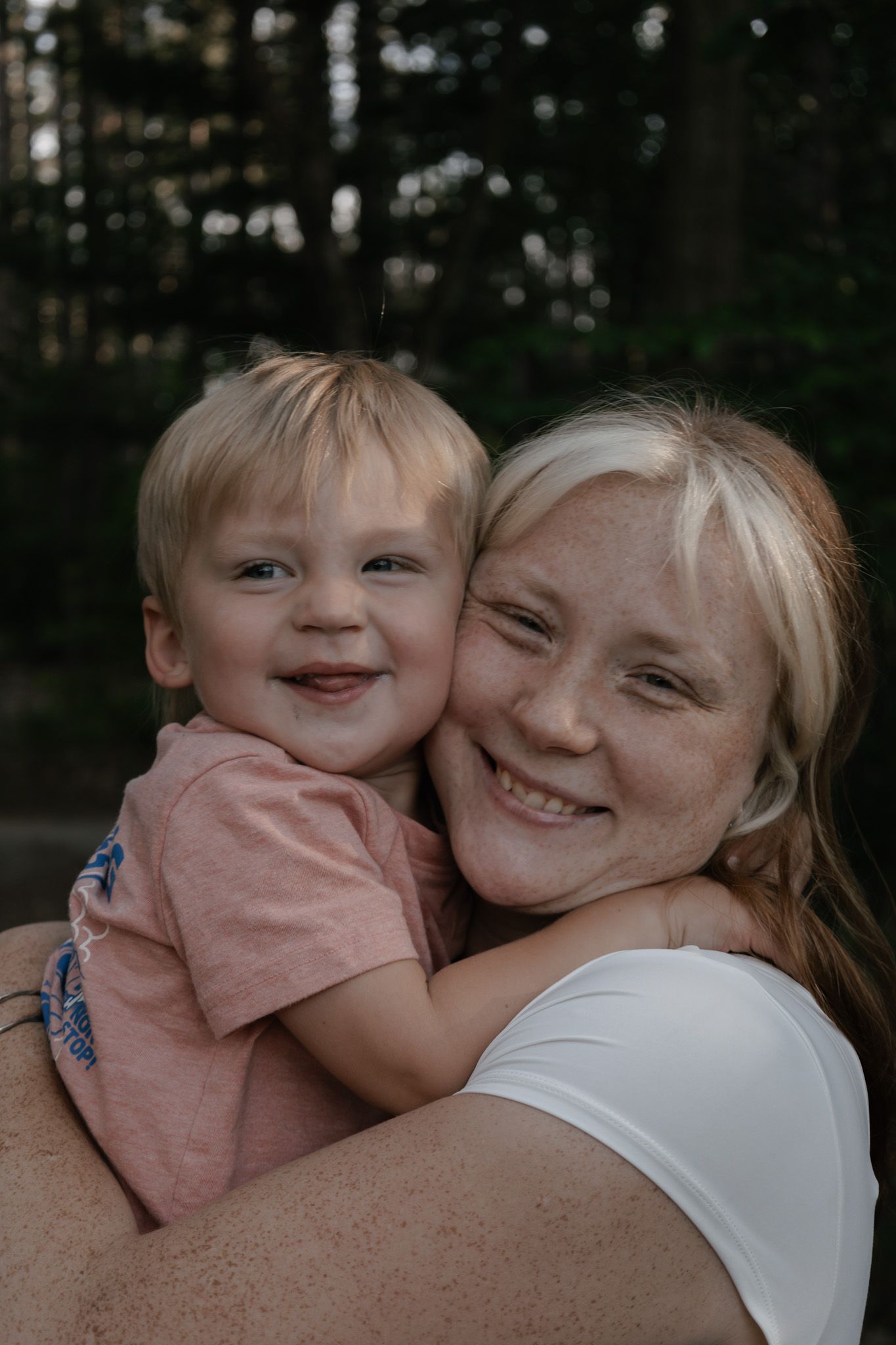 A smiling woman with freckles holds a happy blonde toddler boy outdoors in a forest setting.