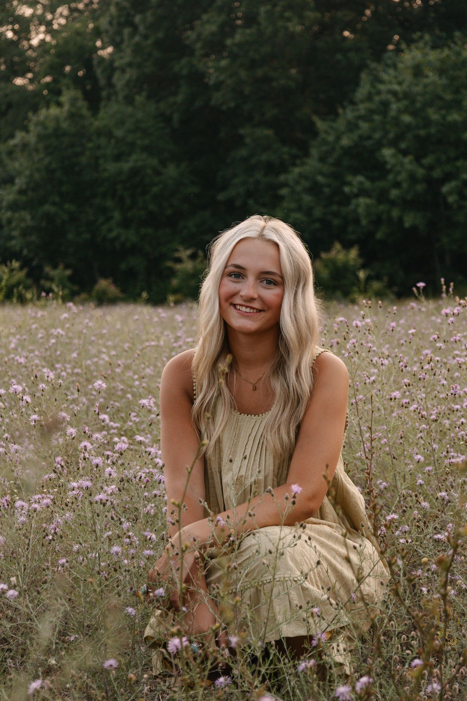 Smiling blonde woman in yellow dress sitting in wildflower meadow with purple flowers and green trees.