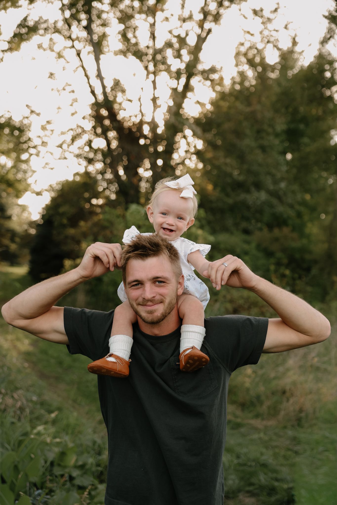 Smiling dad carrying happy baby girl on his shoulders in a lush green outdoor setting.