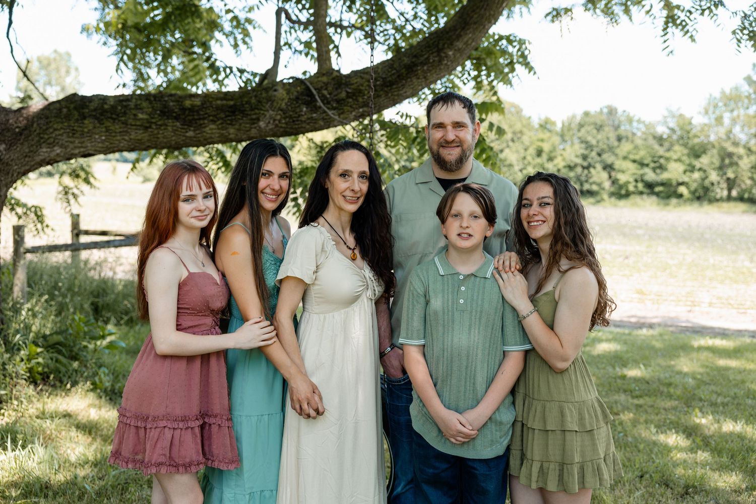 Cheerful extended family of six posing together under a large tree in a lush green outdoor setting.