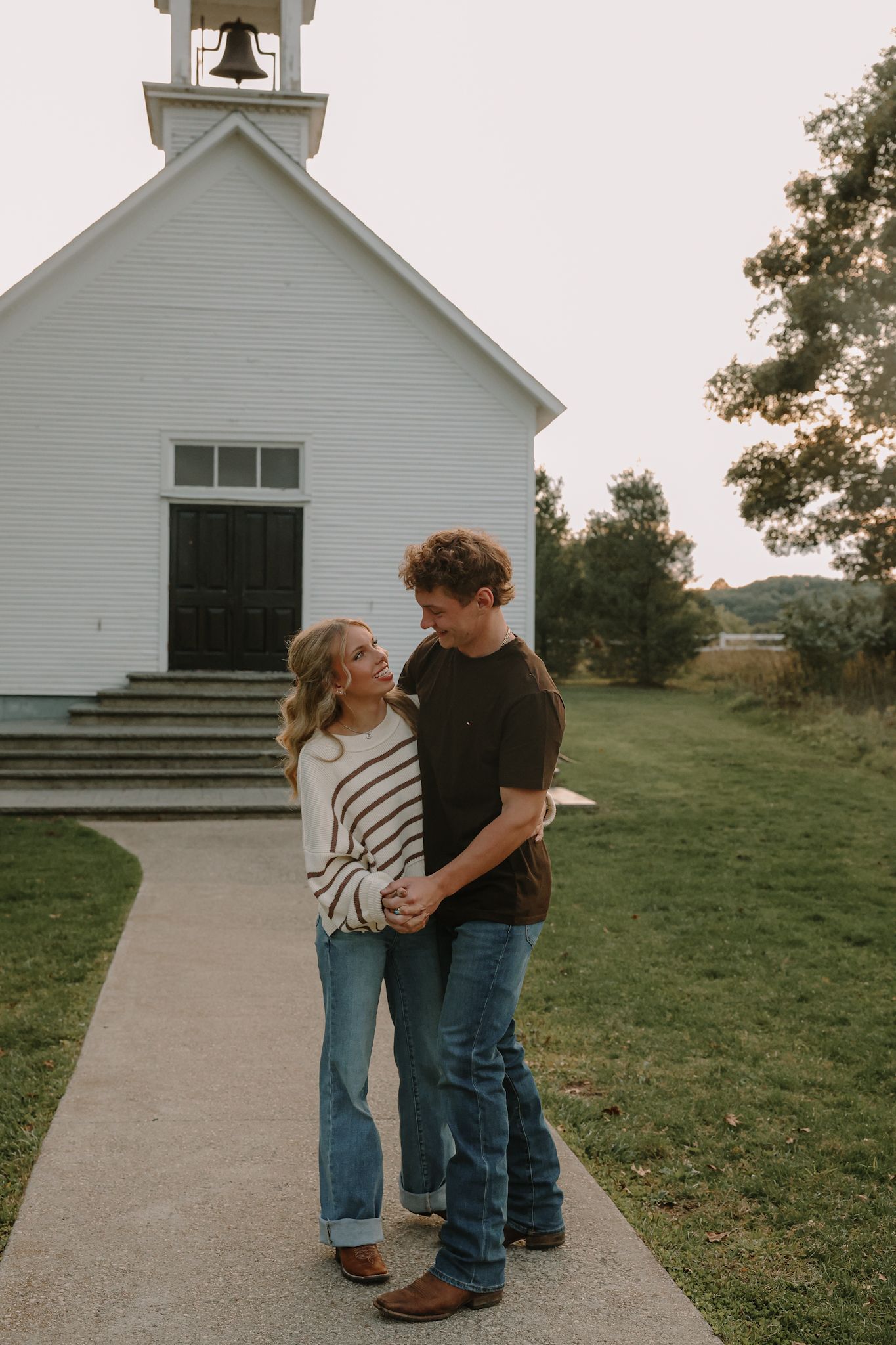 A couple holds hands and smiles at each other on a path outside a classic white country church.