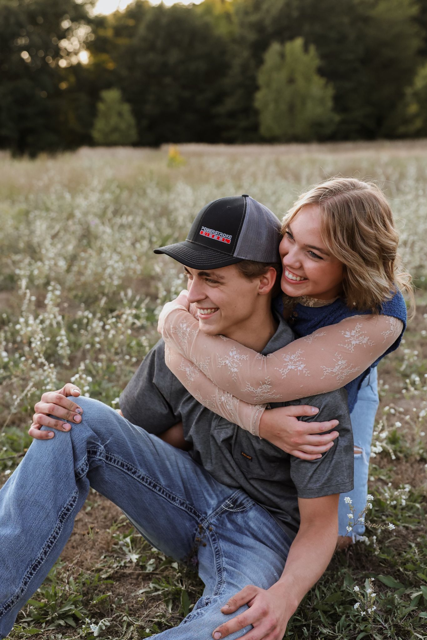 A joyful couple sits together in a blooming field, the woman hugging the man from behind as they both laugh.