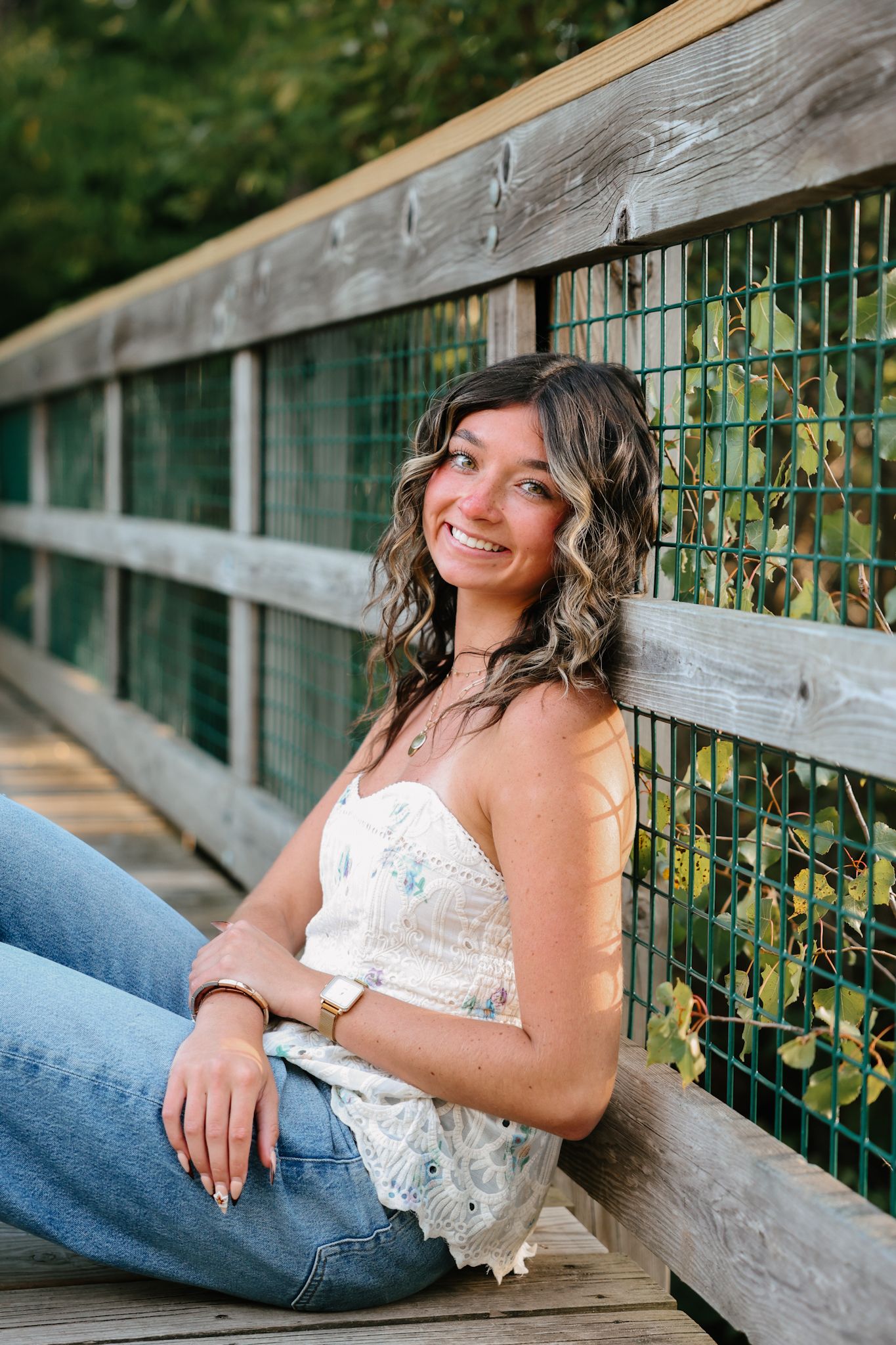 Young woman in floral top and jeans smiling on wooden bridge.