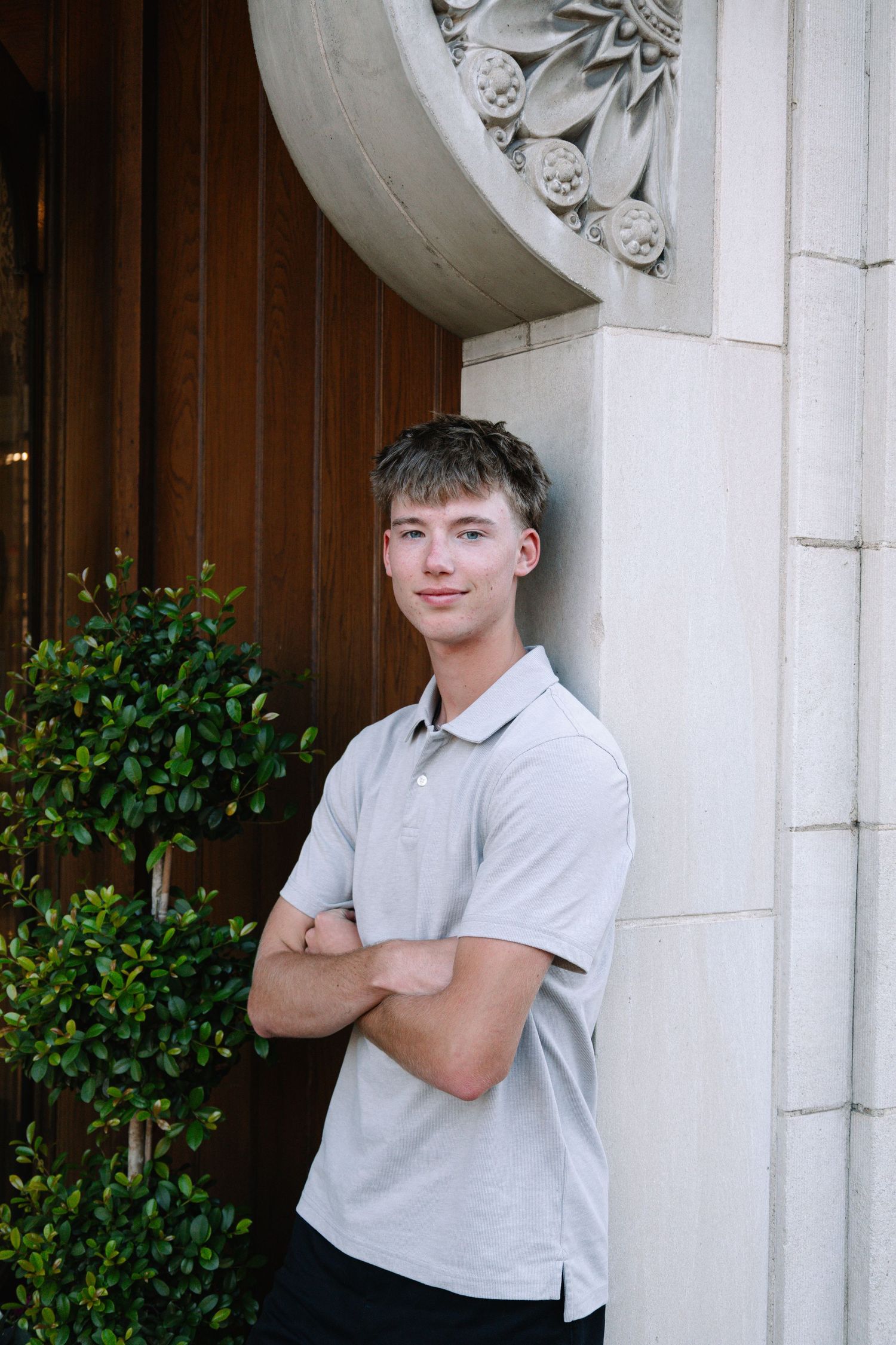 Young man in gray polo shirt stands arms crossed against ornate stone column near wooden door.